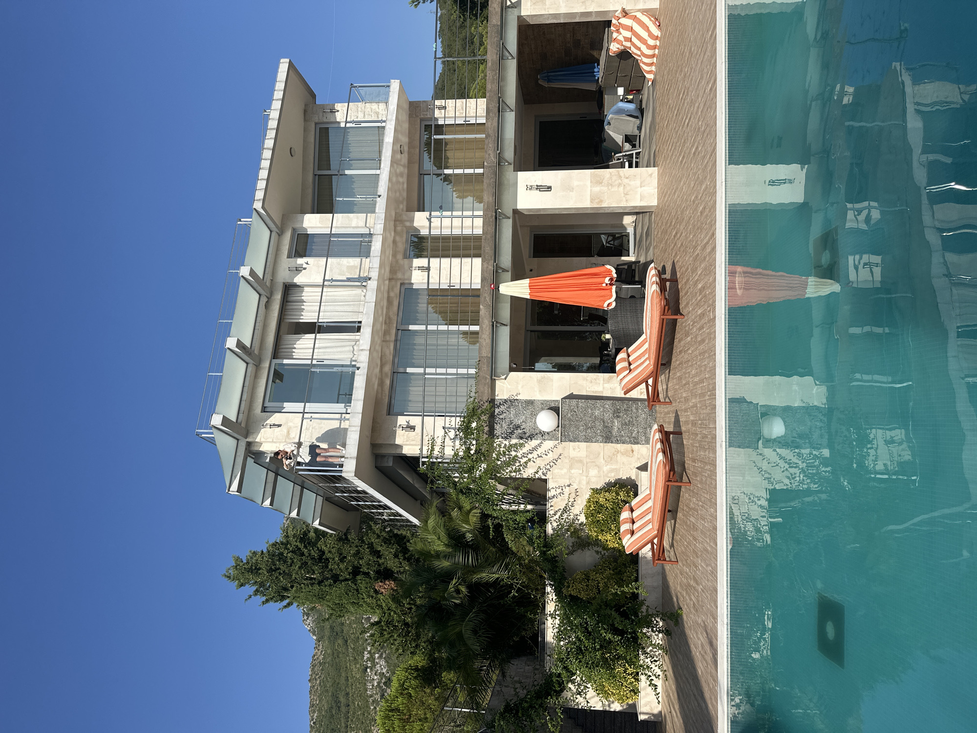 A person is standing on a balcony of a modern multi-story villa in Brca, Montenegro, holding a phone and looking at it. The villa is built with light-colored stone and has large windows and glass railings on its balconies. In front of the villa, there is a swimming pool with clear turquoise water, its surface reflecting the building and the surrounding greenery. On the wooden deck beside the pool, there are two orange and white striped lounge chairs, and an orange umbrella stands partially open near a seating area. The villa is nestled on a hillside with lush green trees and vegetation, with a mountain visible in the background under a clear blue sky.
