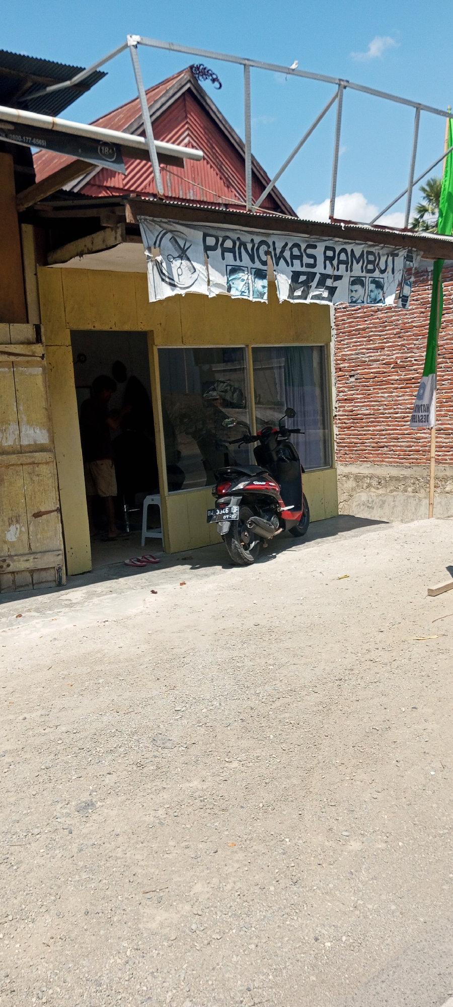 In the city of Lempa, Indonesia, a modest barbershop, Pangkas Rambut 85, is situated. The shop features a yellow facade with a glass storefront, reflecting the bright, clear sky and a nearby palm tree. A banner with scissors and the shop's name hangs above the entrance. Inside, two individuals are visible, one appearing to be a barber attending to a client. A red and black scooter is parked in front of the shop, with a license plate reading "BW 3446 UF 07-22". The area surrounding the shop is unpaved, with a gravelly ground. A brick wall forms the backdrop to the right of the establishment, and a green flag is attached to a wooden pole on the far right.