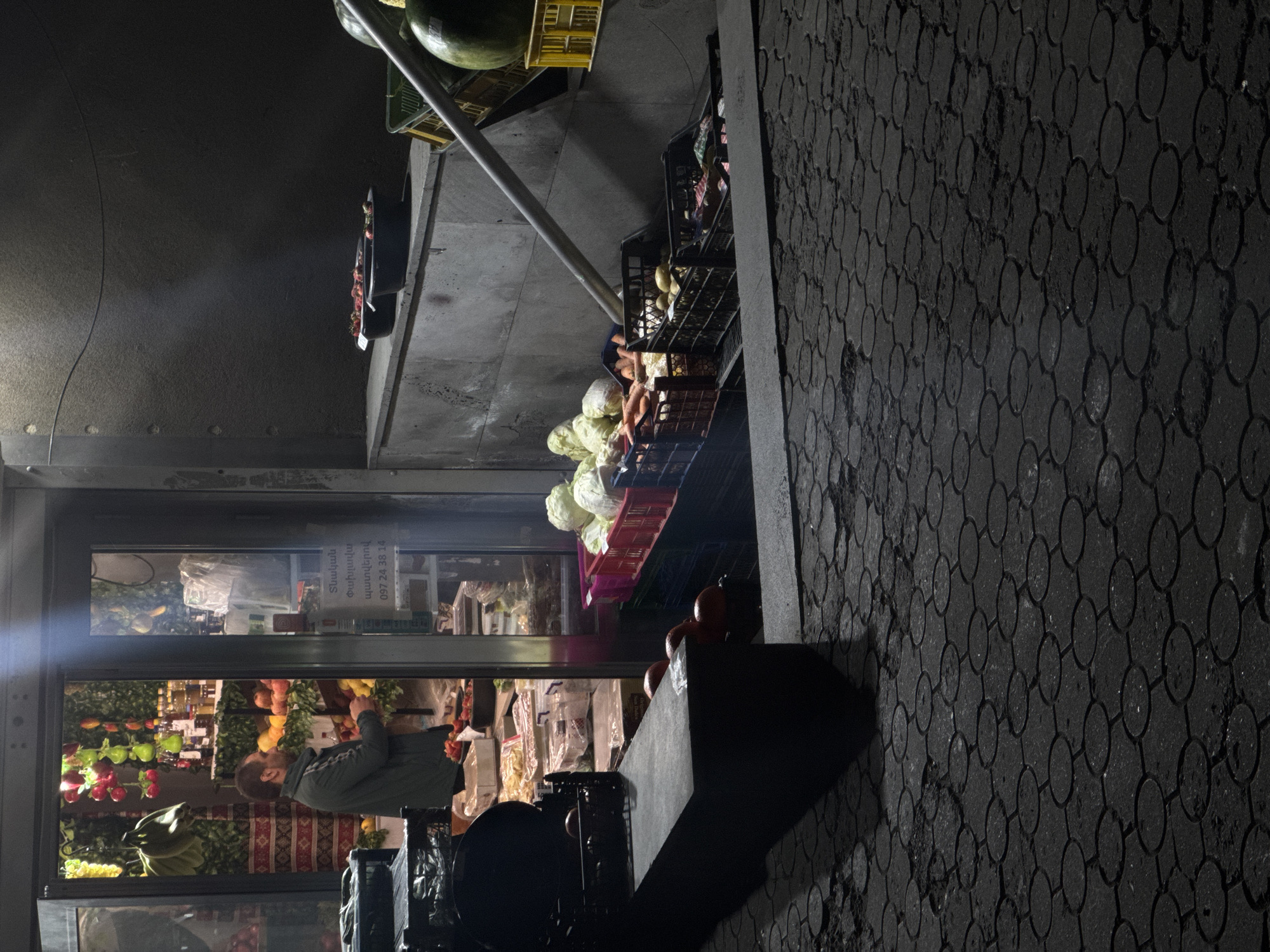 A slightly rotated night shot depicts a Yerevan, Armenia, produce stand. The foreground shows a patterned dark paving stone sidewalk. The produce stand itself is on the left, a walk-in with a man inside appearing to be working. The produce stand window also displays pricing information. Produce is also displayed outside, in crates, including what appears to be cabbage and potatoes. A metal pipe spans the area above the sidewalk.