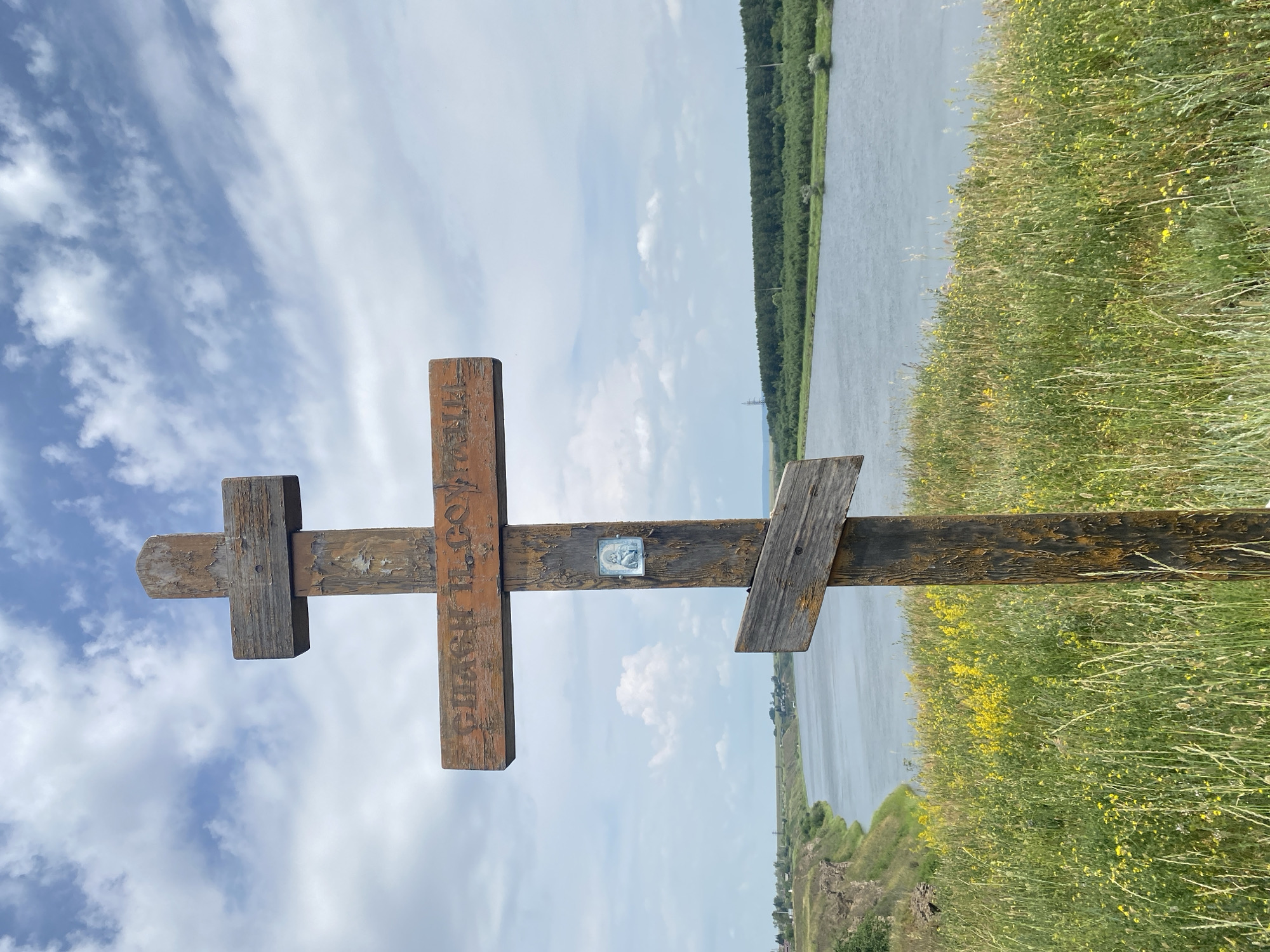The image captures a weathered wooden Christian cross positioned in the foreground, standing on a grassy hillside overlooking a wide, calm river. The cross has an inscription that appears to be in Cyrillic script, reading "СПАСИ И СОХРАНИ" (Save and Protect). A small, blue and white icon, possibly of a religious figure, is affixed to the main post of the cross.

The hillside is covered in lush green grass and dotted with small yellow wildflowers, suggesting a natural, uncultivated setting. The river flows gently in the mid-ground, bordered by a dense line of trees on the opposite bank. The sky above is a mix of blue and white clouds, indicative of a partly cloudy day. In the distance, along the left bank of the river, hints of a settlement or town can be seen, with buildings and what appears to be a rocky, eroded cliff face. Power lines and pylons are visible in the far distance, to the right of the river. The overall impression is one of a peaceful, scenic landscape with a prominent religious marker. The image is taken outdoors during daylight hours.