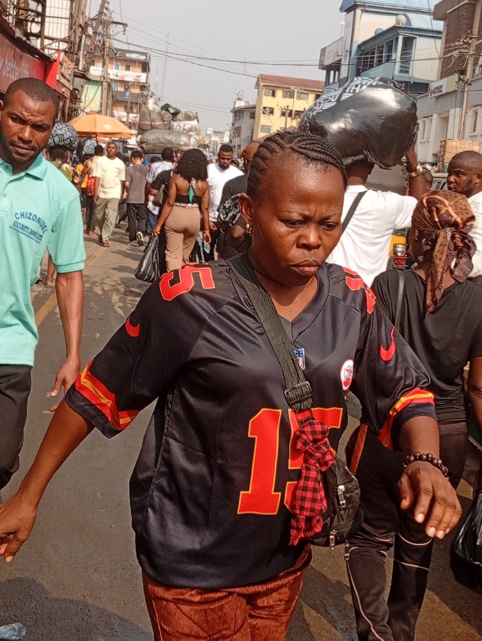 A woman in a black Kansas City Chiefs jersey with the number 15 walks through a crowded street market. A man in a light green shirt walks to her left, while other pedestrians are visible in the background. The street is filled with vendors and people carrying goods. Buildings line the sides of the street under a bright, slightly overcast sky. The woman has a black bag slung across her chest, with a red and black patterned cloth tied to the strap.