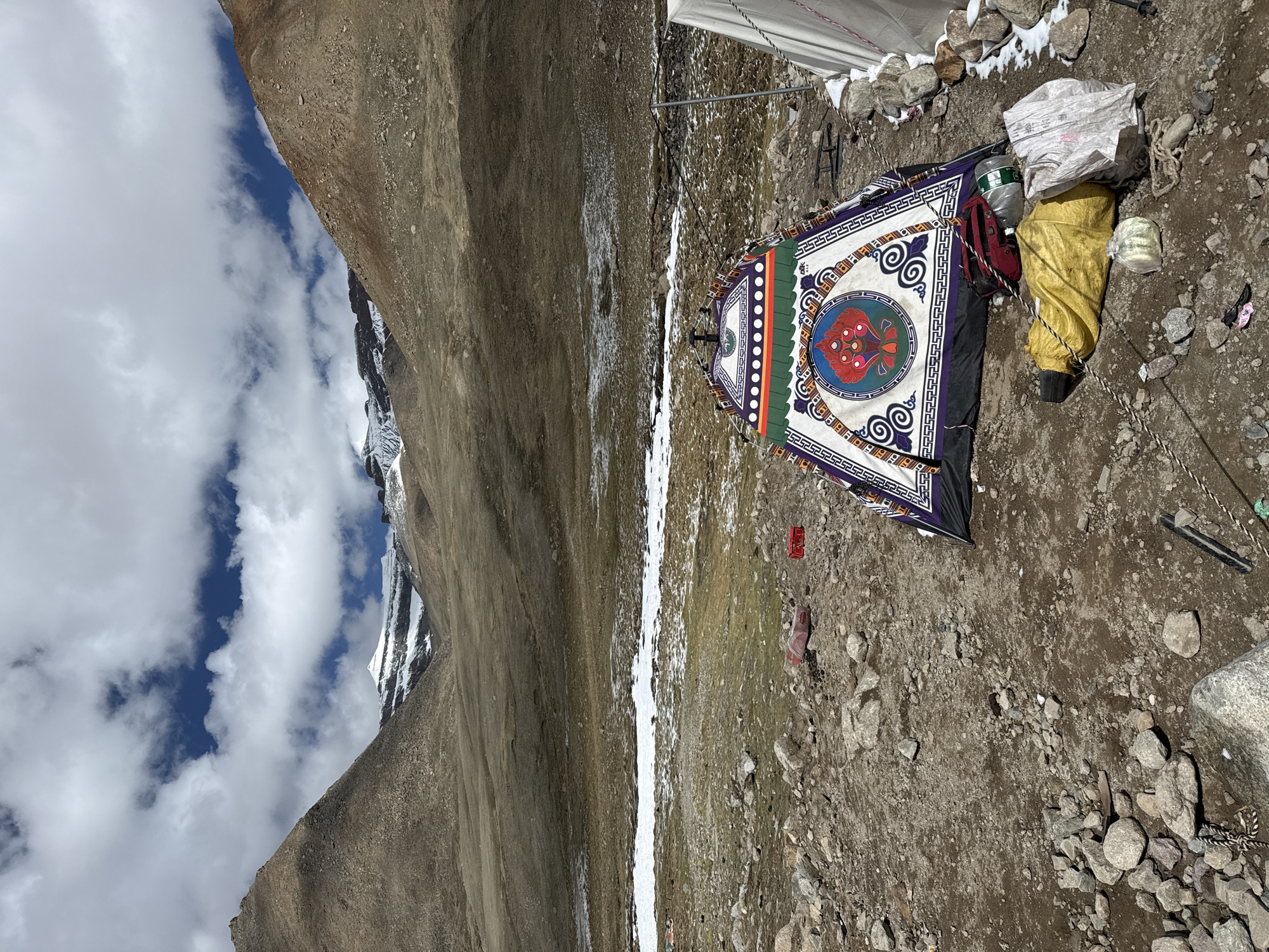 The image depicts a temporary camp or rest stop situated in a rugged, high-altitude mountainous landscape. The ground is uneven and stony, covered with dirt and scattered rocks, along with visible patches of snow, particularly a narrow streak running through the mid-ground.
In the foreground, a brightly colored, rectangular mat or tent flap is laid out, adorned with intricate traditional patterns. It features a geometric border, swirling motifs, and a prominent central circular design in red and blue. Adjacent to this, various items are stacked: a large yellow bag or tarp, a red backpack, a big plastic water bottle, and a white bundled bag. Ropes are visible, likely securing the equipment or a nearby structure.
Behind the foreground items, a portion of a white tent or tarp structure can be seen, supported by poles and tied down with ropes.
The background is dominated by imposing mountains. To the left, there are steep, barren brown slopes, while to the right, snow-capped peaks and a darker, shadowed mountain are visible. The sky is bright and partly cloudy, featuring white cumulus clouds against a clear blue backdrop, indicating daytime, possibly late morning or early afternoon.
No individuals are present in the photograph, but the camp setup suggests recent human activity in this remote environment, possibly related to trekking or temporary settlement. The decorative patterns on the mat suggest a Tibetan or Himalayan cultural influence, fitting the context of Parga town, China. The scene conveys a sense of resilience and remoteness amidst a majestic natural setting.
