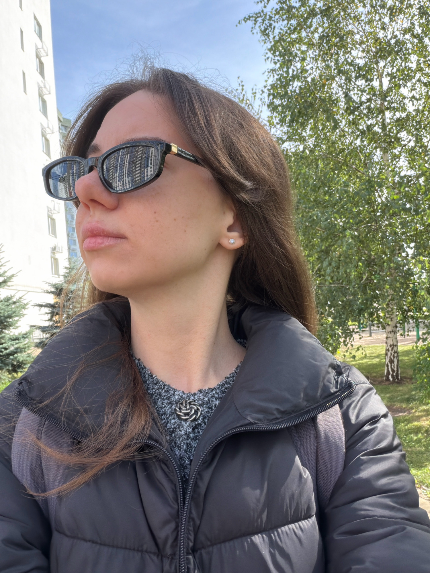 The image shows a close-up, side-profile view of a young woman outdoors, looking to her left. She has brown hair, is wearing dark-rimmed sunglasses, a dark puffer jacket over a grey patterned sweater, and a simple silver pendant necklace. A small, clear stud earring is visible on her left ear. Her backpack is partially visible under her jacket. The background features a tall, white apartment building to the left, trees and greenery to the right, and a clear blue sky above. The lighting suggests it is daytime, likely late morning or early afternoon. The location is identified as a residential area within Kyiv, Ukraine, evidenced by the architecture of the apartment building.