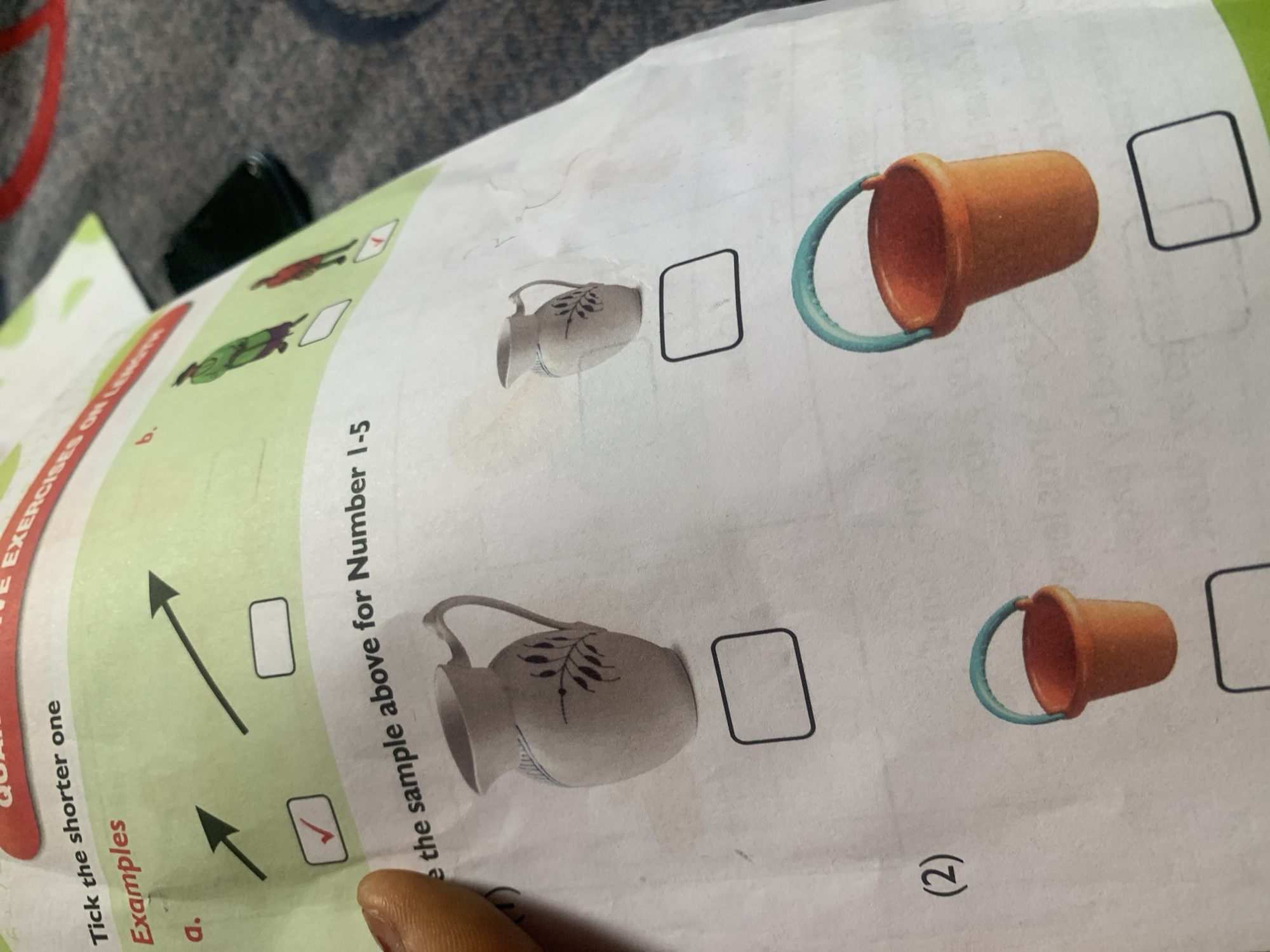 The image shows a page from a workbook or educational material. The top portion displays the text "Tick the shorter one" followed by "Examples." Underneath, there are two illustrated examples labeled "a." and "b." Example "a." shows two arrows, with the shorter one marked with a red checkmark. Example "b." shows two figures of people, one in a green outfit and another in a red outfit. The shorter figure in the red outfit is marked with a red checkmark. Below these examples, the text reads "e the sample above for Number 1-5." The rest of the page shows illustrations of a pitcher and a bucket, each with an empty box below it, presumably for the student to tick the shorter one. The image is taken from an overhead perspective, capturing part of a table or surface in the background. There is no information within the image that relates to the city of Jauro Yinu, Nigeria.