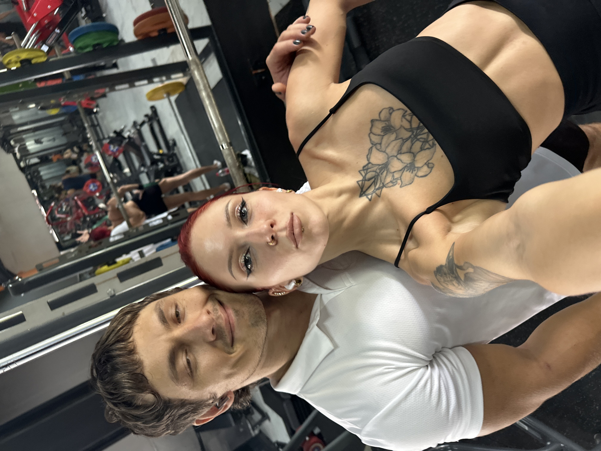 A selfie captures two individuals inside a gym located in North Kuta, Indonesia. The woman, with striking red hair, a nose ring, and a lip piercing, holds the camera. She wears a black sports bra or top, revealing two prominent tattoos: a large floral design on her left chest/shoulder and a winged, bird-like figure on her right arm. Her expression is neutral.
Leaning his head against her shoulder/neck, the man, with brown hair and a slight smile, wears a white polo shirt.
The background is dominated by large mirrors that reflect the interior of the gym, showcasing various pieces of exercise equipment. Visible in the reflections are colorful weight plates (yellow, blue, green, orange, red), barbells, and exercise racks. Several other individuals can be seen in the mirrored background, engaged in their workouts, suggesting an active gym environment. The image conveys a casual, affectionate moment between the two subjects in a fitness setting. No explicit text is visible in the photograph.