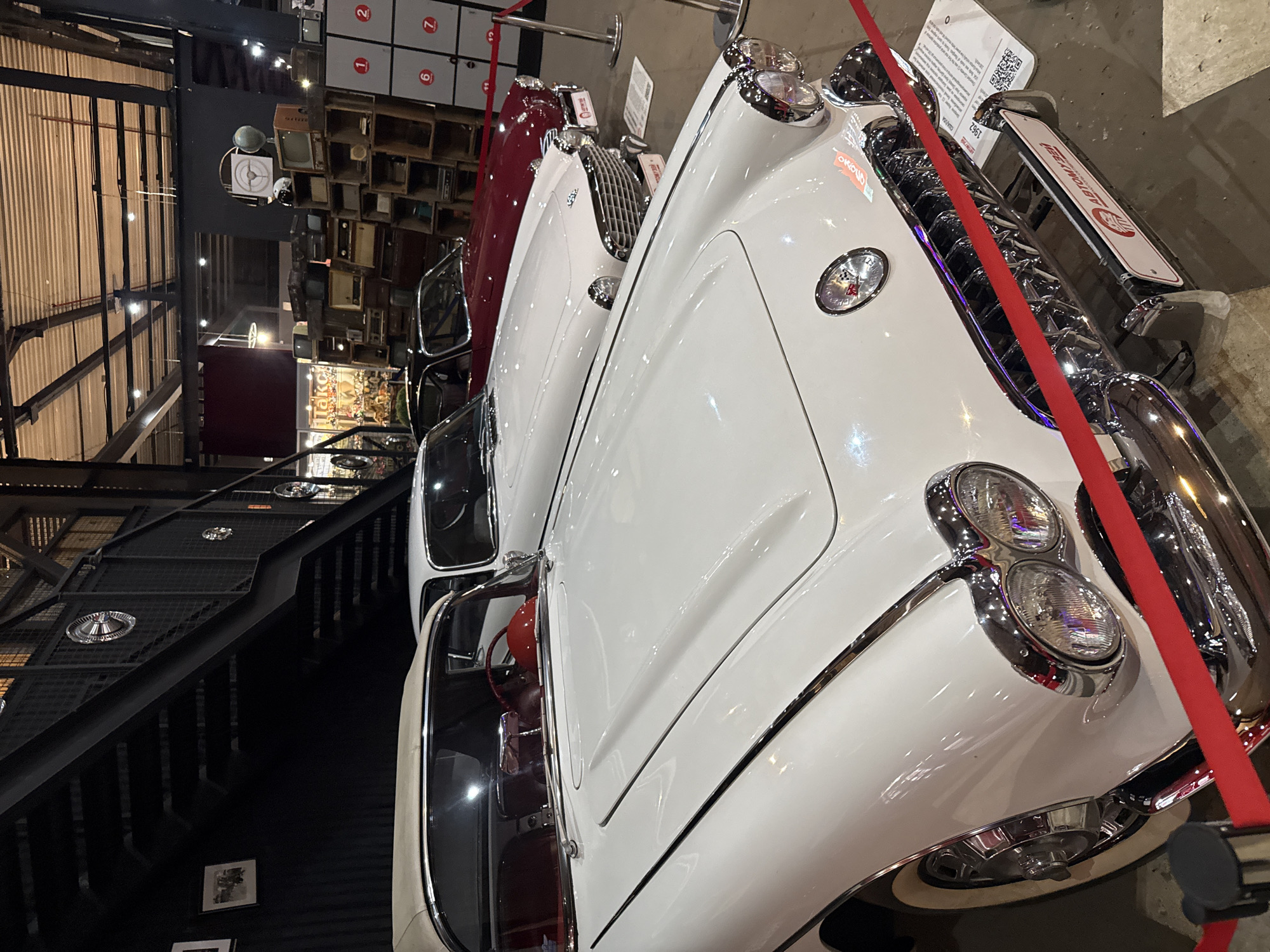 The image shows a museum interior with several vintage cars on display. The primary focus is on the front of a white 1962 Chevrolet Corvette Stingray, its chrome grille and dual headlights prominent. To the right of the Corvette, a red classic car is partially visible. In the background, a dark metal staircase and industrial-style ceiling structure are visible, suggesting an industrial or warehouse setting adapted for exhibition. Further in the background, shelves are stacked with vintage electronics like televisions and radios. The cars are separated from viewers by a red velvet rope barrier. Signs are attached to the cars, one with the text "ABTOMYZEЙ" and a QR code. The overall impression is a well-lit exhibition space showcasing vintage automotive and electronic artifacts. The sign "ABTOMYZEЙ" translates to "Automuseum", indicating the location is an automotive museum. Although the exact city location is not explicitly stated in the image, the context of the prompt specifies Moscow, Russia.