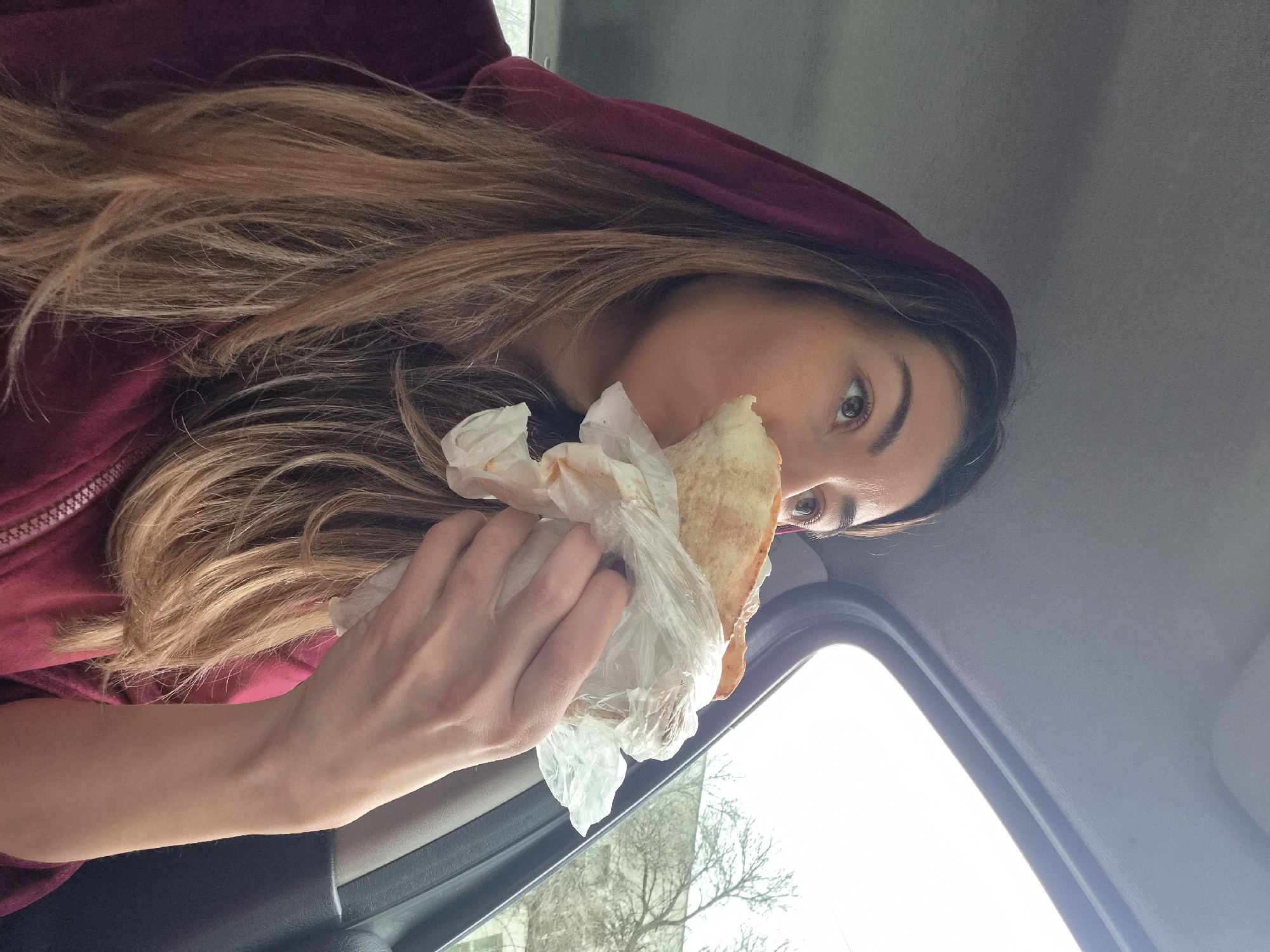 A woman with long, light brown hair and wearing a maroon hooded sweatshirt is seated inside a car. She is holding and biting into a sandwich wrapped in white paper. The interior of the car is visible, including the door frame and window. Outside the window, bare trees and a light-colored building can be seen. The location cannot be determined from the image.