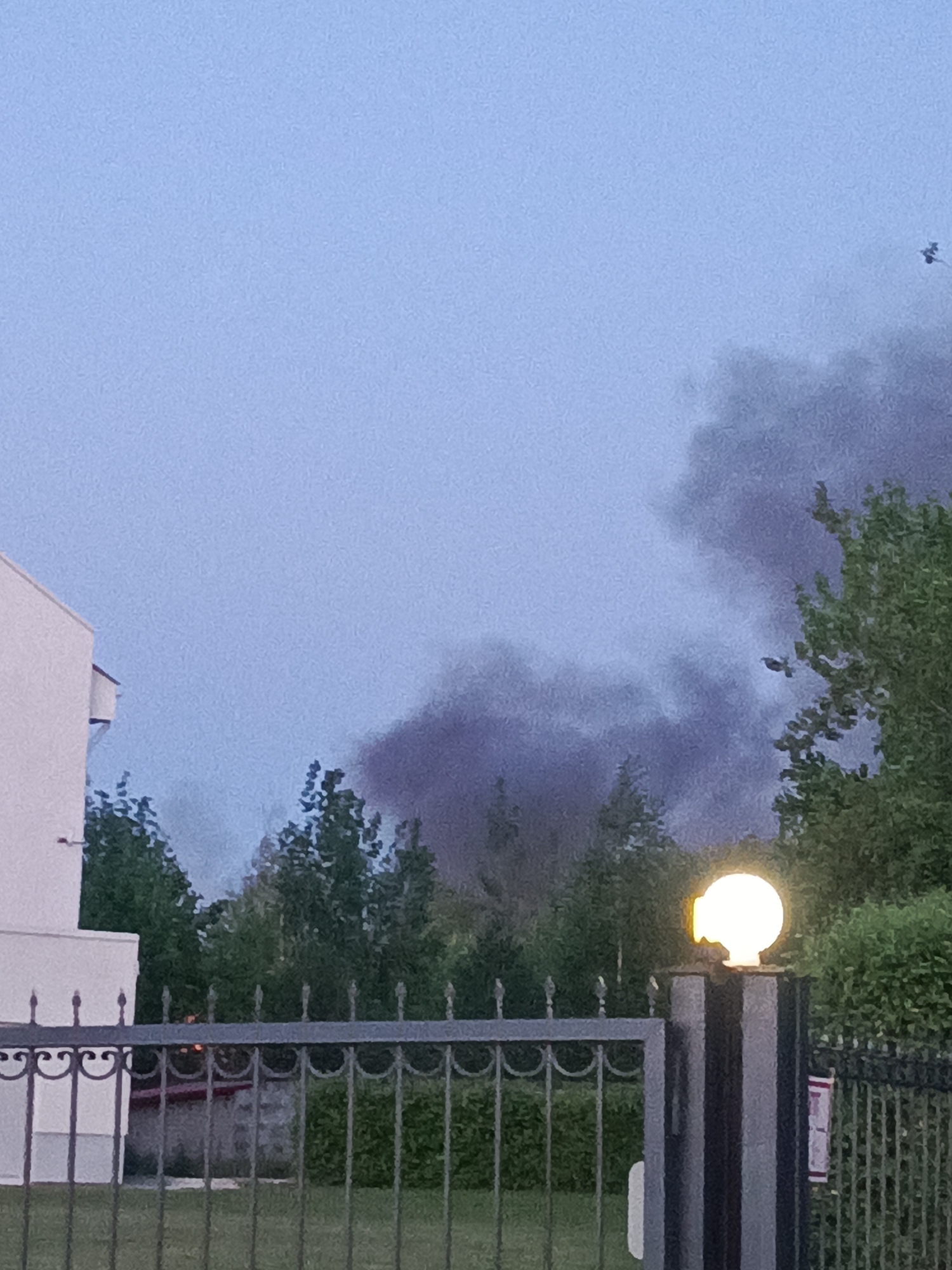 The image depicts a scene in what appears to be a residential area. In the foreground is a metal fence with a decorative design. Behind the fence is a lawn and a building to the left. In the background, there is a line of trees, and rising above the trees is dark smoke. A street lamp is illuminated on the right side of the image, attached to a gate post. In the upper right corner, a small object that could be a bird is visible. The location is Chaikovsky, Russia. There are no people visible in the image.