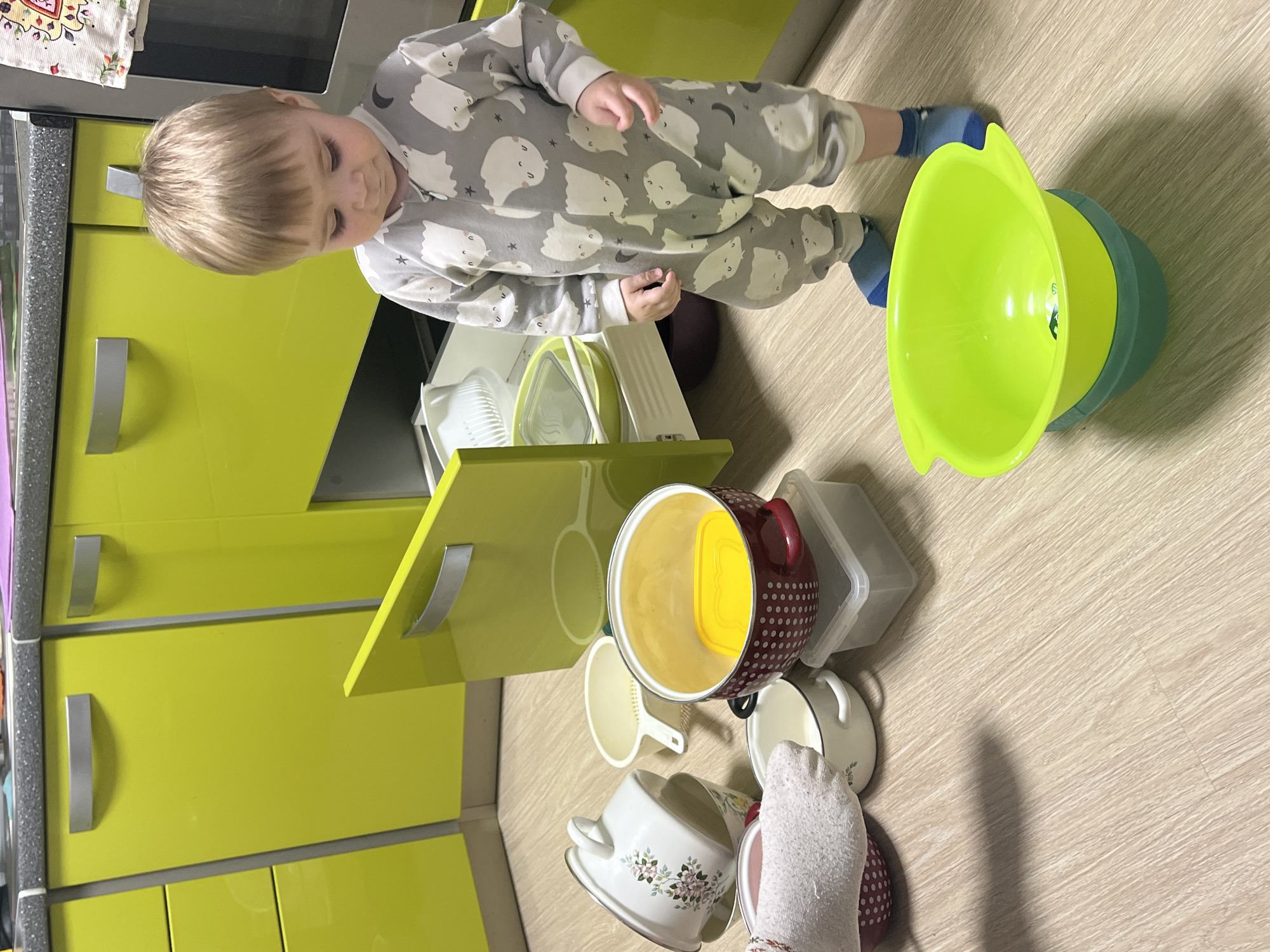 A young child with light blond hair, dressed in a grey sleepsuit adorned with white ghost-like figures and stars, and blue socks, stands in a kitchen. The child is positioned centrally in the frame, looking down with a focused expression at an array of kitchen items scattered on the light wooden laminate floor.
The scattered items include a prominent bright lime green mixing bowl, a maroon pot with white polka dots, a white pot with a delicate floral design, several white ceramic mugs, a white plastic colander, and clear plastic storage containers.
The kitchen features modern, vibrant lime green cabinets with sleek silver handles. A dark, speckled countertop is visible on the left, topped with a patterned cloth. One of the cabinet doors on the lower level is slightly ajar, suggesting the source of some of the scattered items. The lighting is bright and even, typical of an indoor scene during the day. The image captures a moment of childhood exploration or play within a home setting in Perm, Russia. No visible text is discernible in the image.
