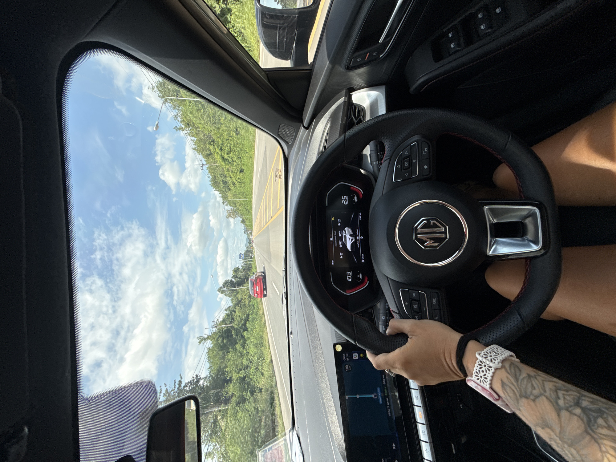 The image shows a first-person perspective from inside a car driving on a road. The driver's hands are visible on the steering wheel. The dashboard displays the car's speed at 103 km/h and a navigation screen showing a route. The scene outside the car includes a tree-lined road, power lines, and a blue sky with white clouds. A red car is visible ahead on the road. The location is identified as Don Sak, Thailand.