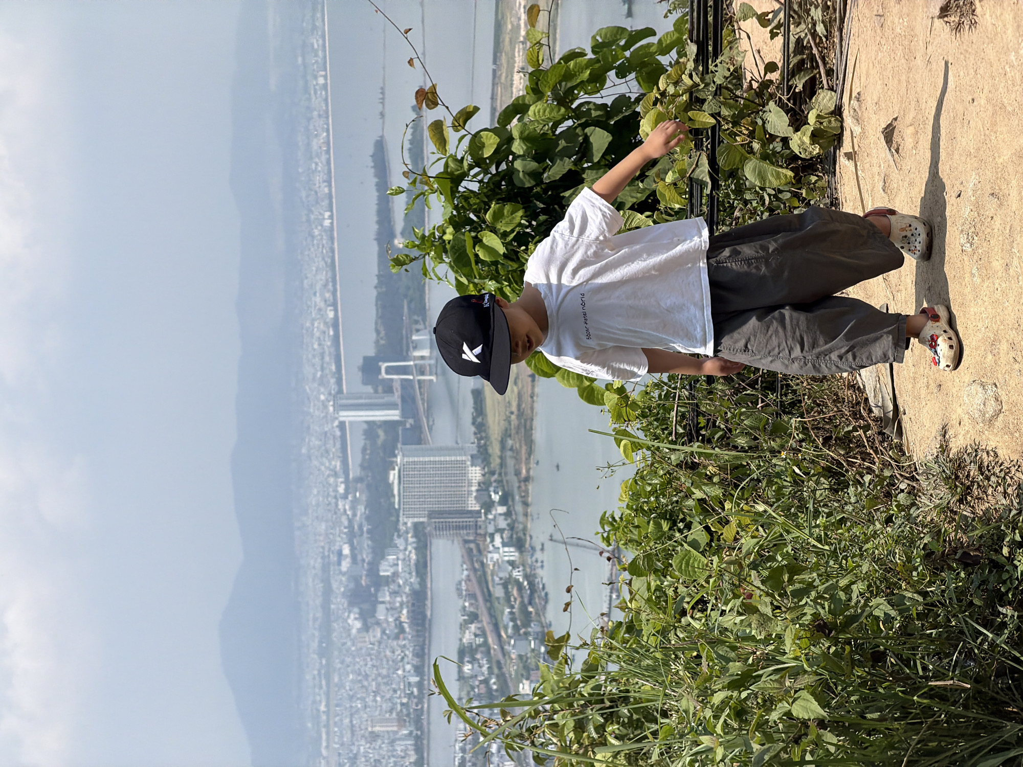 A young person stands in front of dense foliage on a hillside, overlooking a cityscape and waterway in Đà Nẵng, Vietnam.
The individual is wearing a white short-sleeved shirt, olive-green pants, a black baseball cap with a white logo, and slip-on shoes with decorative embellishments. They are positioned near the edge of a concrete surface, partially obscured by overgrown vegetation. Their hand is extended towards the foliage.
In the background, a wide river or bay separates the foreground hill from the cityscape, which includes numerous buildings, a bridge, and a hazy mountainous region. The sky is overcast, contributing to a diffused light throughout the scene.