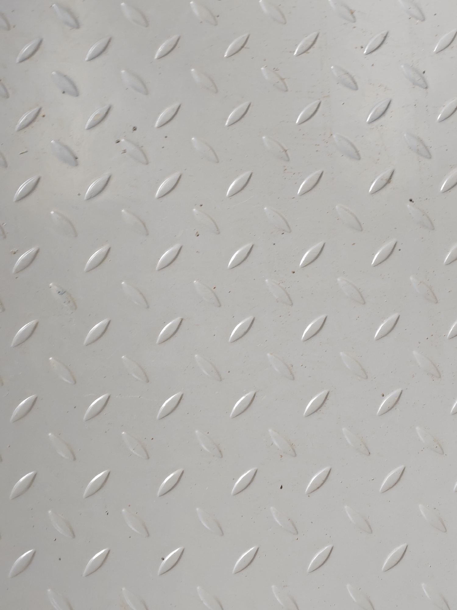 The image is a close-up, overhead shot of a light gray metal surface with a raised diamond pattern. The pattern consists of rows of elongated diamond shapes, each with a smooth, slightly reflective surface. The texture is consistent across the entire frame, with subtle variations in lighting creating a sense of depth and highlighting the raised elements. There are a few very small dark specks scattered across the surface, possibly dust or debris. The surface appears to be an industrial or utilitarian material, such as diamond plate flooring or paneling.
The image is a generic, close-up view of a textured metal surface and does not contain any specific visual cues that would allow for its localization to Saint Petersburg, Russia, or any other specific city.