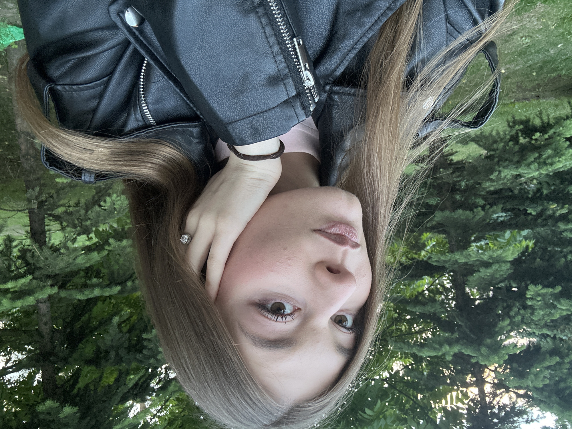 The image, originally rotated 90 degrees counter-clockwise, features a young woman in an outdoor setting, looking directly at the camera. She has long, straight light brown hair that frames her face, and light brown eyes. Her lips are naturally colored or have a light gloss, and faint freckles are visible on her skin. She is dressed in a black leather-look jacket with a prominent silver zipper, worn over a light pink top. Her left hand is raised, with fingers gently resting against her cheek and jawline. A silver ring with a large clear stone adorns her left ring finger, and a dark brown hair tie or bracelet is on her left wrist.
The background consists of dense green foliage, dominated by coniferous trees, indicating a park or forested environment. Sunlight filters through the leaves, suggesting it is daytime. The woman's expression is direct and focused. The overall scene conveys a natural and tranquil ambiance. The location context provided is Kazan, Russia.