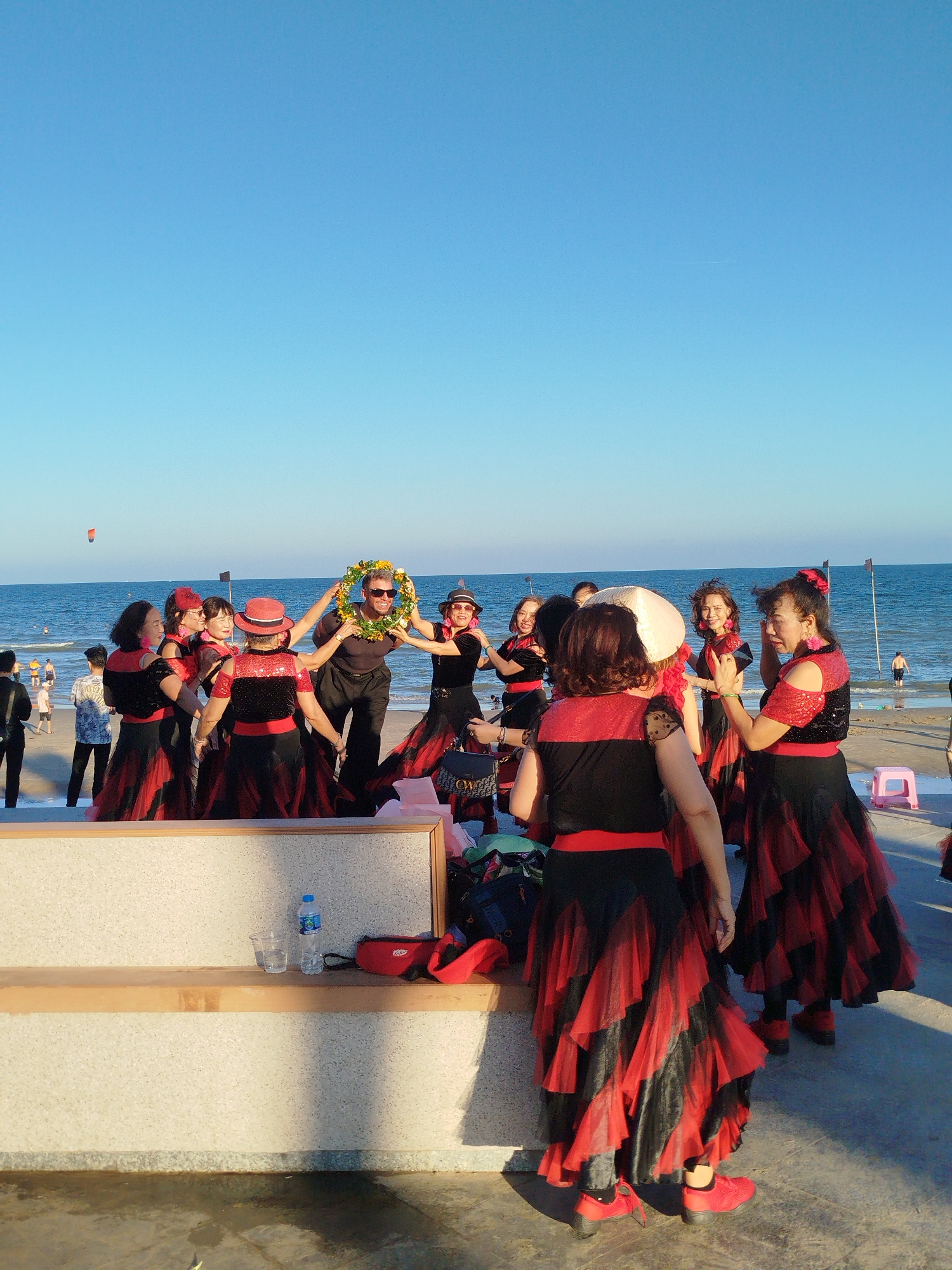 A group of people is gathered on a beach, with a clear blue sky and ocean as the backdrop. The group consists of women wearing matching red and black dresses, and a man standing in the center of the group holding a floral wreath over his head. There is a bench in the foreground with a bottle of water and other items on it. People can be seen walking on the beach in the background. A kite is visible in the sky. The photograph was taken at Ho Chi Minh City, Vietnam.