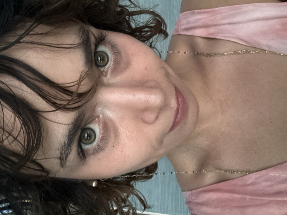 A close-up, slightly angled shot features a person with dark, curly hair and striking light green eyes. The individual appears to be wearing a light pink top with shoulder straps, and a delicate, beaded necklace graces the neck. The focus is on the face, highlighting the eyebrows, nose, and lips, conveying a sense of intimacy and vulnerability.