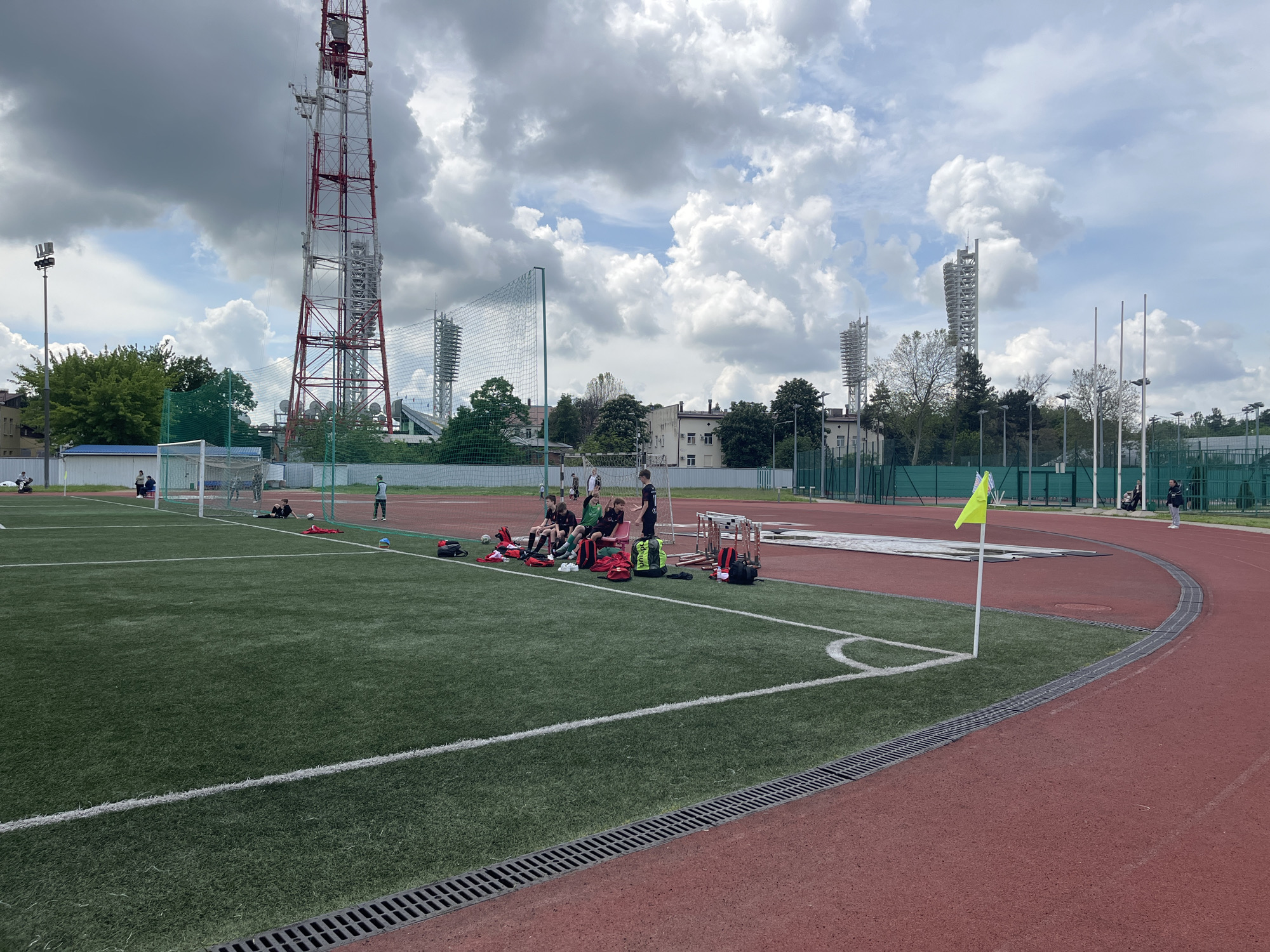 The image shows a soccer field with a running track in Krasnodar, Russia. In the background are tall communications towers and stadium lighting. A group of young people are sitting on the sidelines of the field, near some equipment and bags. One individual is standing nearby. A soccer goal is visible on the far side of the field. A yellow flag marks the corner of the field near the running track. The sky is overcast with large clouds. The running track is red. A drainage ditch runs along the edge of the field.