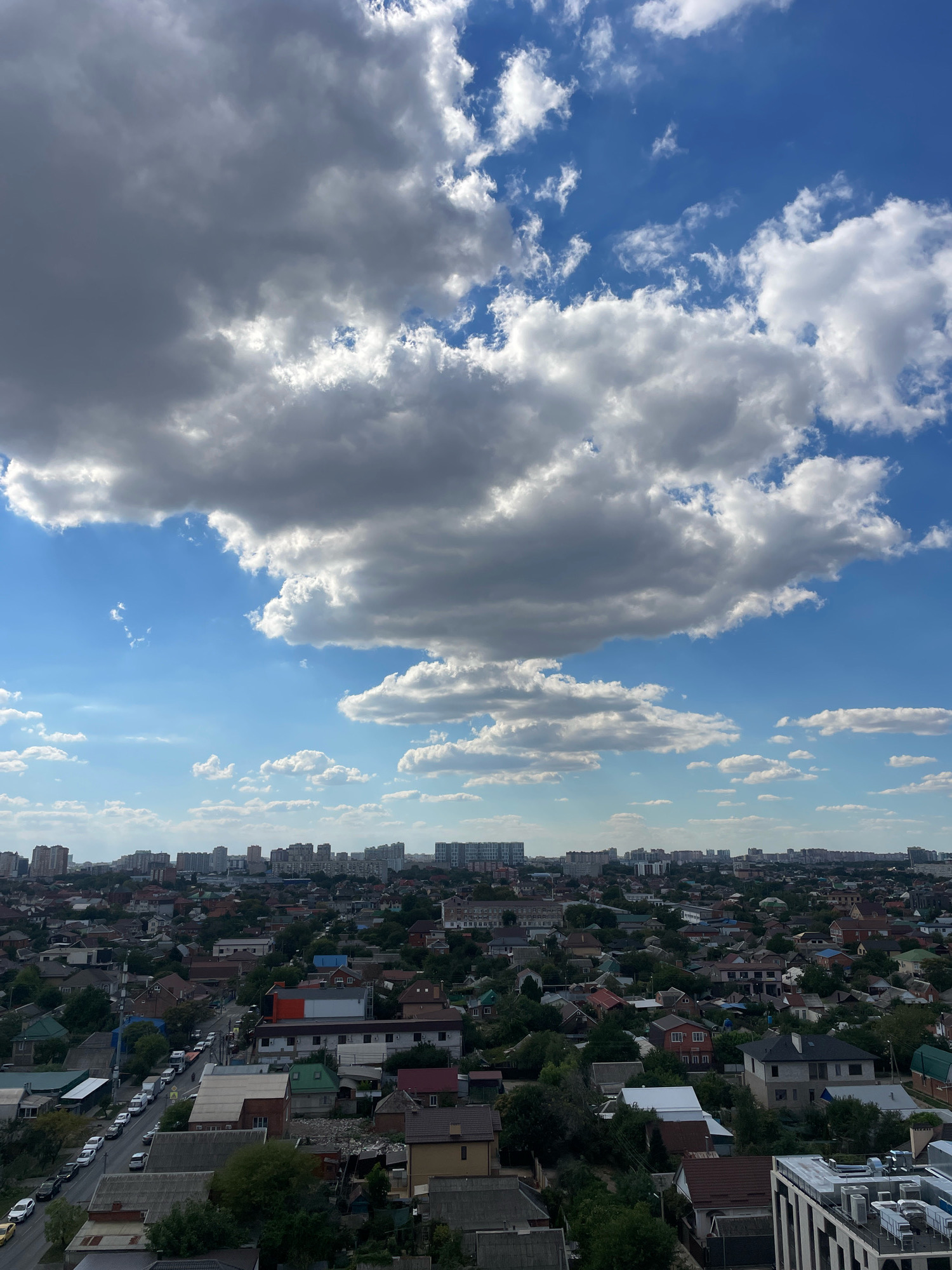 This is a high-angle, vertical shot of a suburban area within the city of Krasnodar, Russia, under a partly cloudy sky. The foreground features the rooftops of numerous houses, predominantly one and two stories high, with varied roofing materials such as shingles and metal. Some houses have visible yards with trees and vegetation. A street runs through the middle of the frame, lined with parked cars, including sedans and vans. Further in the distance, a denser collection of apartment buildings forms the city skyline, with some modern high-rises among older structures. The sky is a prominent element, with large, dramatic cumulonimbus clouds casting shadows over parts of the city, contrasting with patches of bright blue sky. The overall impression is of a residential neighborhood on a day with dynamic weather. There are no visible people or animals in the image.