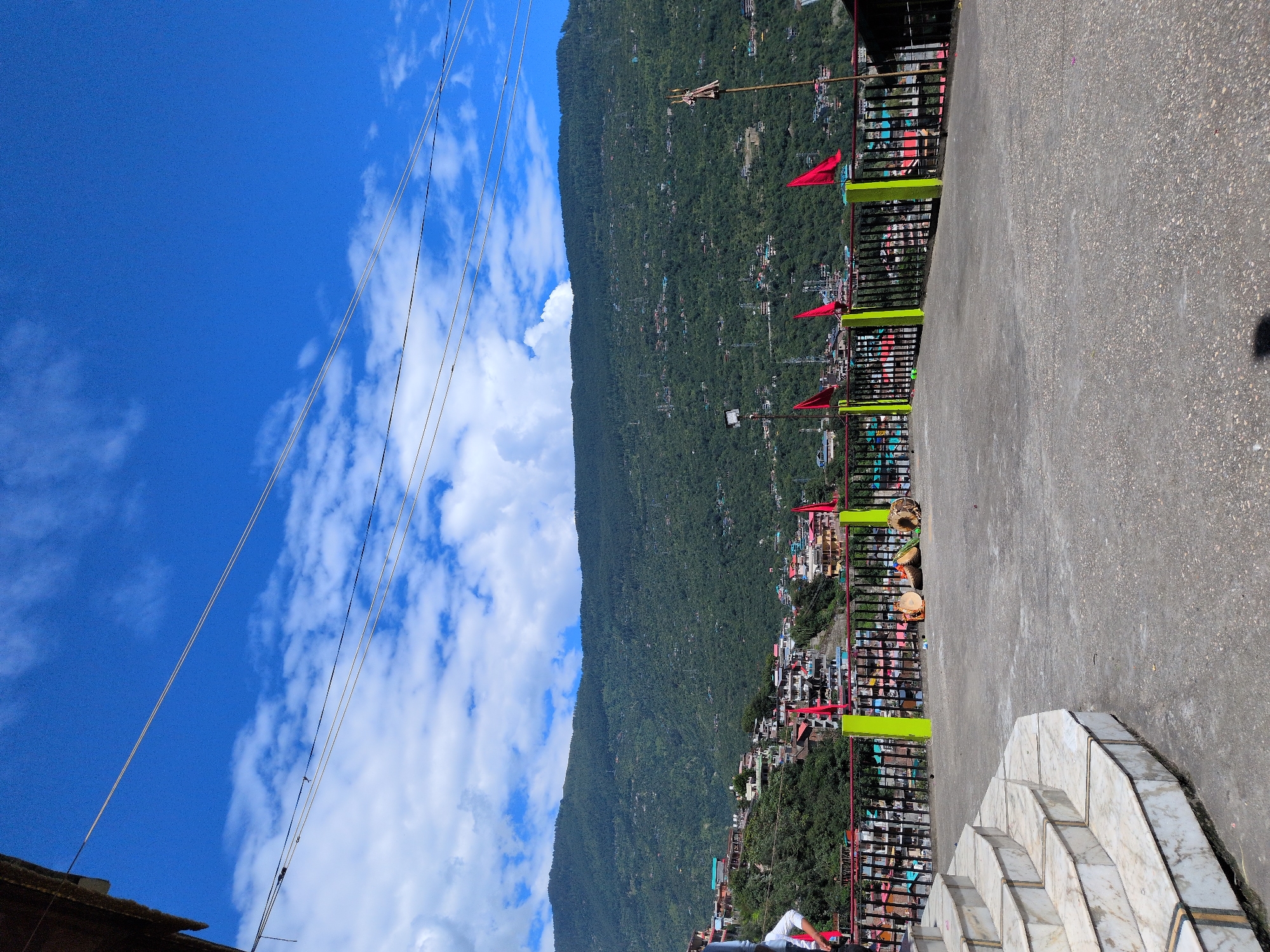 In Kullu, India, a paved rooftop platform overlooks a town nestled on a forested mountainside. The platform features a white marble staircase on the left, leading up to the viewpoint. A black metal railing with bright green posts borders the platform, separating it from the town below. Several red flags are affixed to the railing, and two large drums are placed on the ground near the railing. In the background, the town’s buildings, with colorful roofs, are clustered on the steep slopes of the verdant mountain. The sky above is a vibrant blue, dotted with white, fluffy clouds. Two individuals are visible on the far left, one standing and the other partially visible from the waist down. Electrical wires crisscross the sky.