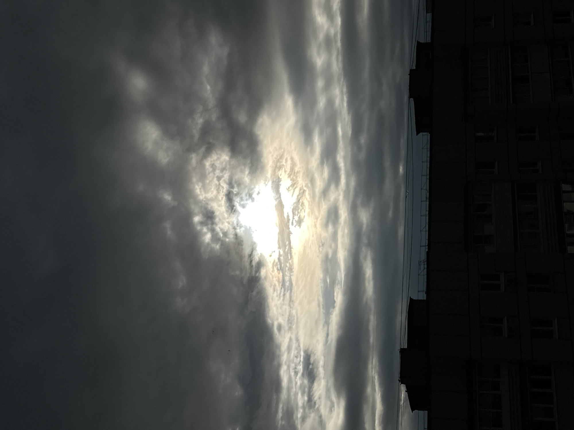 The image captures a dramatic sky with the sun partially obscured by dark, ominous clouds. The sun's rays burst through a gap in the clouds, creating a bright, ethereal glow with golden and white hues. The clouds are varied in texture, with some appearing dense and heavy, while others are lighter and more wispy.
To the right of the sky, a tall, dark building occupies the frame, its silhouette sharp against the dramatic sky. The building appears to be a multi-story structure with numerous windows. Some of the windows are dark, suggesting they are unlit or have blinds drawn, while others might reflect the dim light. The building's architecture is somewhat monolithic and imposing.
The orientation of the image is landscape, but the scene is presented vertically, making the sky appear to stretch upwards and the building to run along one side. This vertical orientation adds to the dramatic and perhaps unsettling mood of the scene.
The time of day appears to be either early morning or late afternoon, given the low angle of the sun and the quality of light. The weather is clearly overcast, with the possibility of an approaching storm or the tail end of one, given the darkness of the clouds. The presence of the building suggests an urban or semi-urban setting.
There are no visible people or text in the image. The overall mood conveyed is one of awe and perhaps a touch of foreboding, with the powerful natural display of the sky juxtaposed against the man-made structure. The image evokes a sense of the sublime, where nature's forces are on full display.
