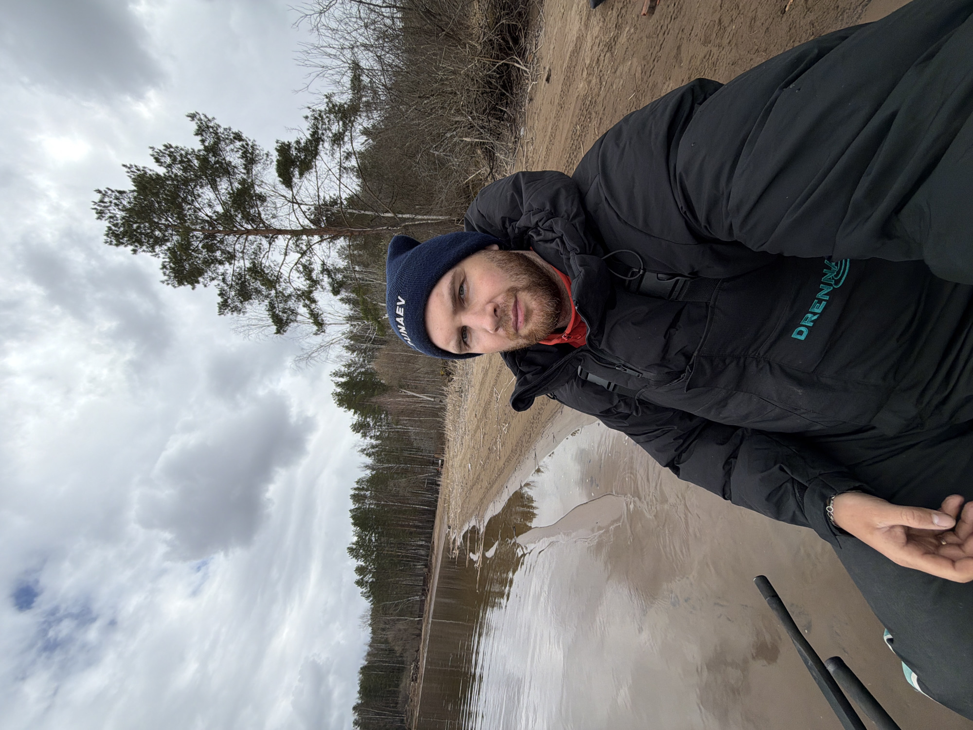 A man is seated at a shoreline in Chaykovsky, Russia. He wears a dark blue beanie and a black winter jacket. He faces the camera with a slight smile. The terrain includes sand, water, trees, and a cloudy sky. A few fishing rods are visible to the right of the man.