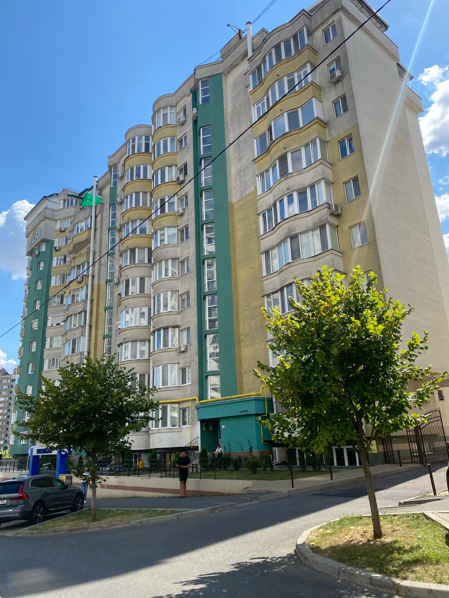 A low-angle, full-shot photograph captures a modern, multi-story residential building situated in Chișinău, Moldova, under a bright, partly cloudy blue sky. The building, characterized by its curved facades and a mix of pale yellow and green exterior paint, features numerous balconies with expansive windows. A prominent green flag is visible on the upper left section of the building. In the foreground, a paved road curves to the right, with a sidewalk and a grassy area bordering it. A dark gray Volvo SUV is parked on the left side of the road. A young person in dark shorts and a t-shirt stands on the sidewalk, looking down at their phone. A mature, leafy green tree is positioned in the mid-ground, casting shadows on the pavement. In the background, other buildings and fences are visible, suggesting an urban residential environment.