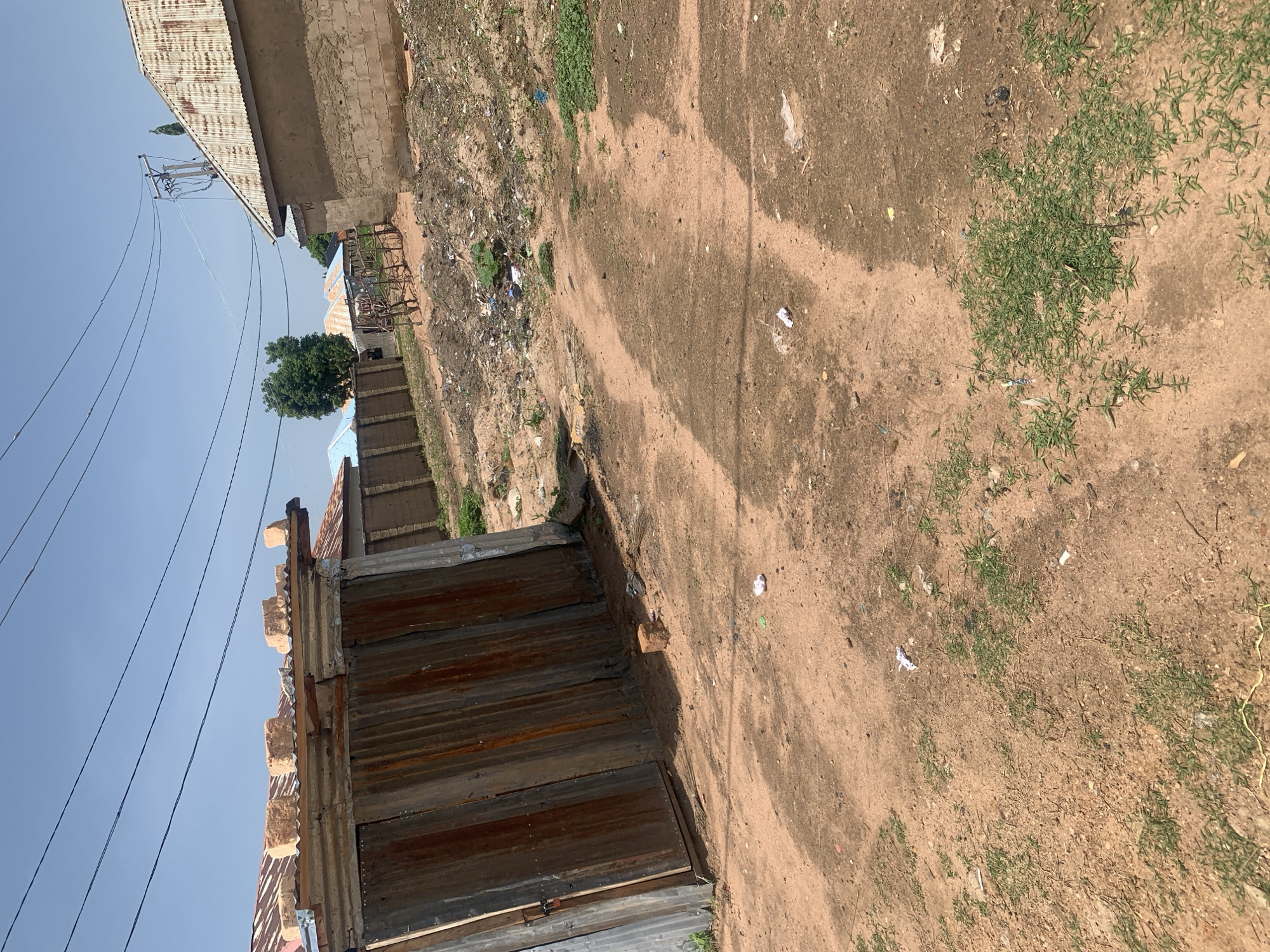 The image depicts a dusty, outdoor scene in the city of Jauro Yinu, Nigeria. In the foreground, a dirt path is visible, with scattered green grass and some litter. To the left, a weathered corrugated metal gate stands partially open, revealing its rusty texture. Beyond the gate, more corrugated metal structures and buildings with rusted tin roofs are visible. In the background, various buildings with mud brick walls and tin roofs are spread across the landscape. Power lines crisscross the sky, supported by wooden poles. A small, rickety wooden cart is also present, along with a pile of debris and a single brick on the ground. The overall impression is one of a somewhat neglected, rural or semi-urban environment under a bright, clear sky.