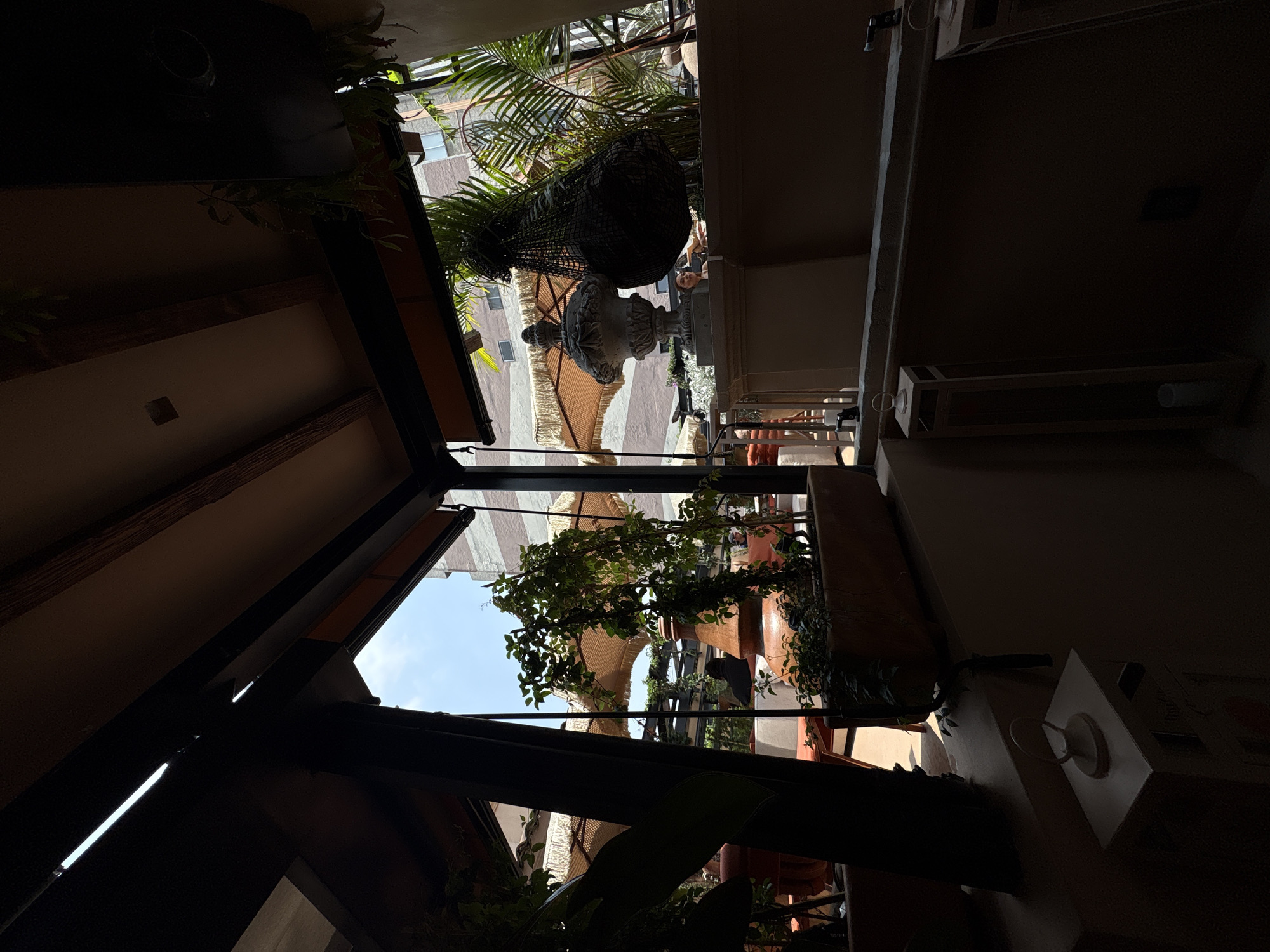 The image depicts an outdoor seating area, likely a restaurant or cafe, with a view of buildings in Mexico City, Mexico. The foreground is dominated by dark shadows, with structural beams and a partial view of a planter box with greenery. Sunlight streams in from the right, illuminating a large stone urn and a black netted bag hanging with some contents. Beyond this, patio umbrellas with fringe trim are visible, along with seating areas and other patrons. The architecture of the surrounding buildings is visible in the background. The scene suggests a bright, sunny day with a relaxed ambiance.