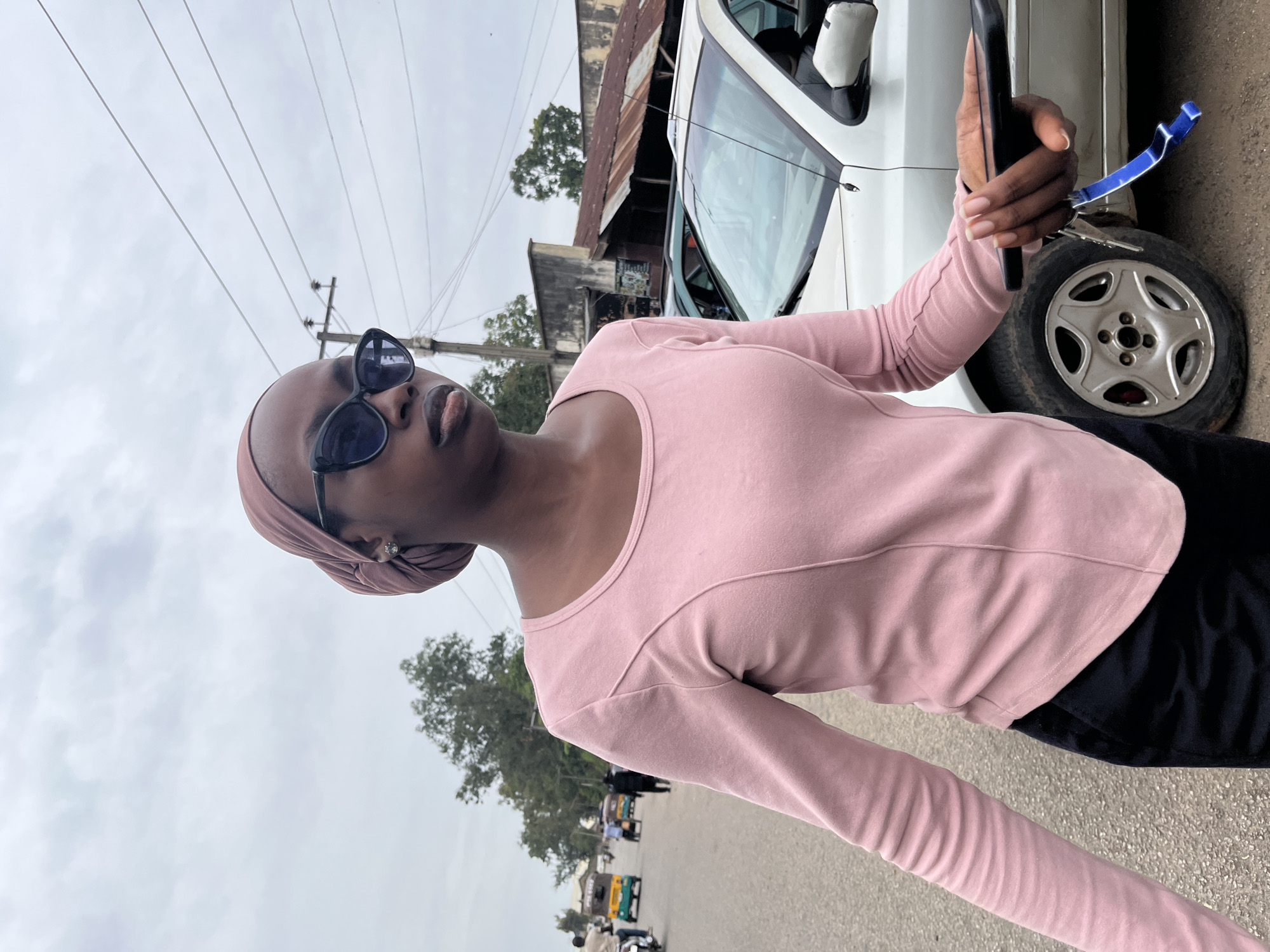 A person is taking a selfie outdoors, holding a dark-colored smartphone in their right hand. They are wearing a light pink long-sleeved top and a light brown headscarf. Dark sunglasses are perched on their face. The person is standing next to a white vehicle. The background shows a street scene with buildings, trees, and other vehicles, including what appears to be a tuk-tuk. Overhead, power lines crisscross the cloudy sky, suggesting an overcast day. The overall impression is casual and everyday. No visible text is present in the image. The location context provided is Jos, Nigeria.