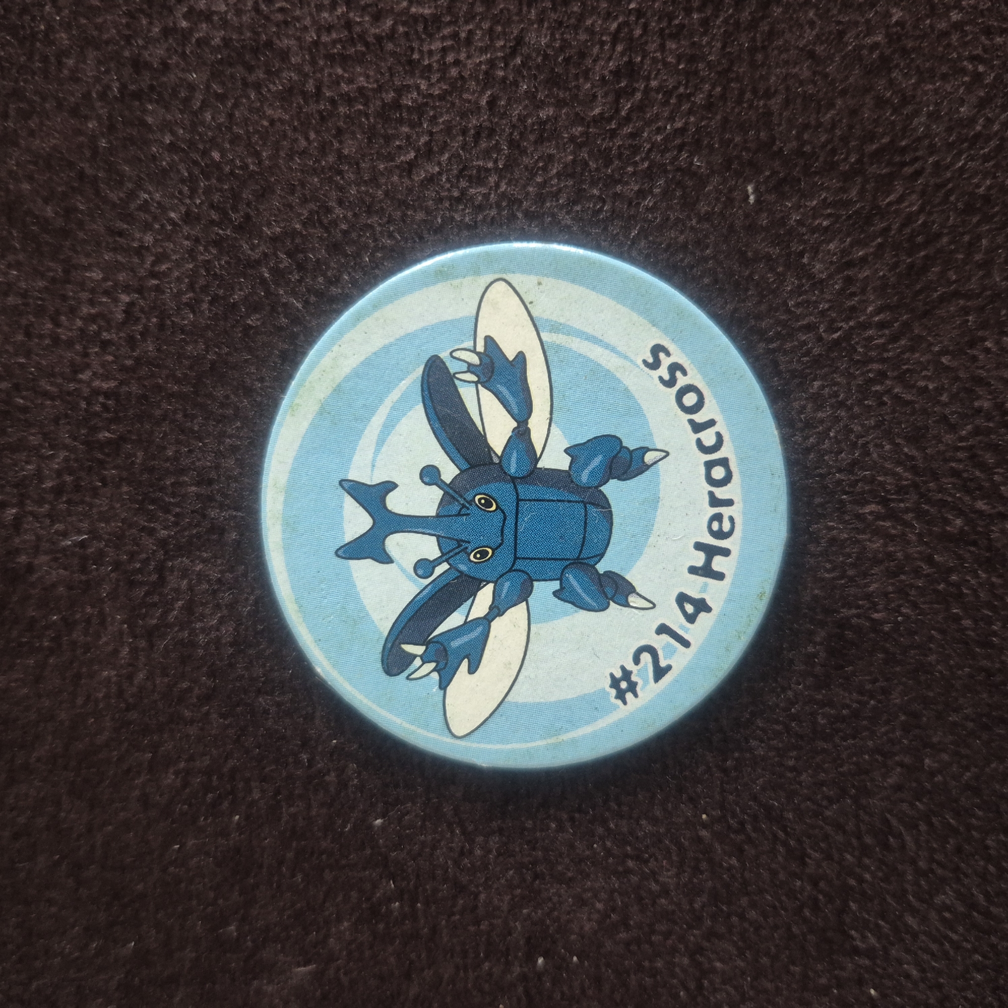 The image shows a close-up, overhead shot of a round, light blue token with a cartoon drawing of the Pokémon Heracross in the center. The token is placed on a dark brown, textured surface that appears to be fabric. The Heracross is depicted as a blue beetle with large wings, horns, and claws, facing forward. The number "#214" and the name "Heracross" are printed in dark blue text along the bottom edge of the token. The background of the token features light blue concentric circles. The image has a shallow depth of field, with the token in sharp focus and the surrounding surface slightly blurred. There is no identifiable location in the city of Kępno, Poland visible in the image.