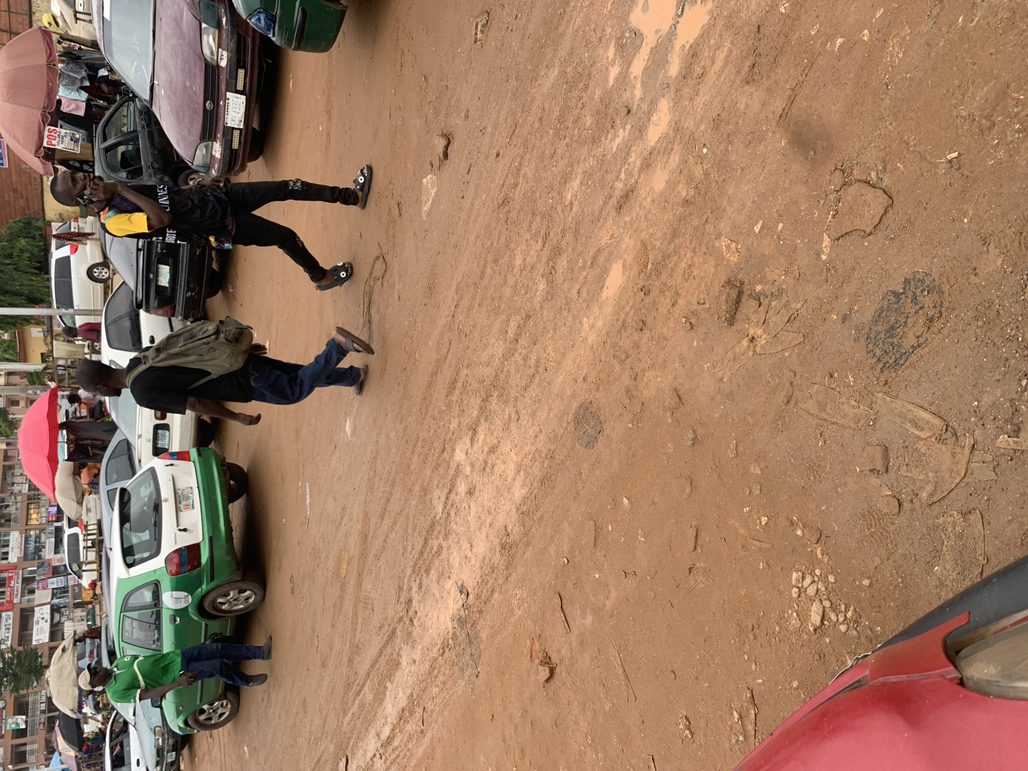 In a street scene in Abuja, Nigeria, a man in a green shirt and blue jeans walks alongside a green and white hatchback car. Another man, wearing a black shirt with colorful accents and black pants, is talking on his phone while walking. The ground is a mix of reddish-brown dirt and mud, with some puddles reflecting the overcast sky. Several other cars are visible in the background, along with market stalls and buildings. The overall impression is of a busy, unpaved urban environment.