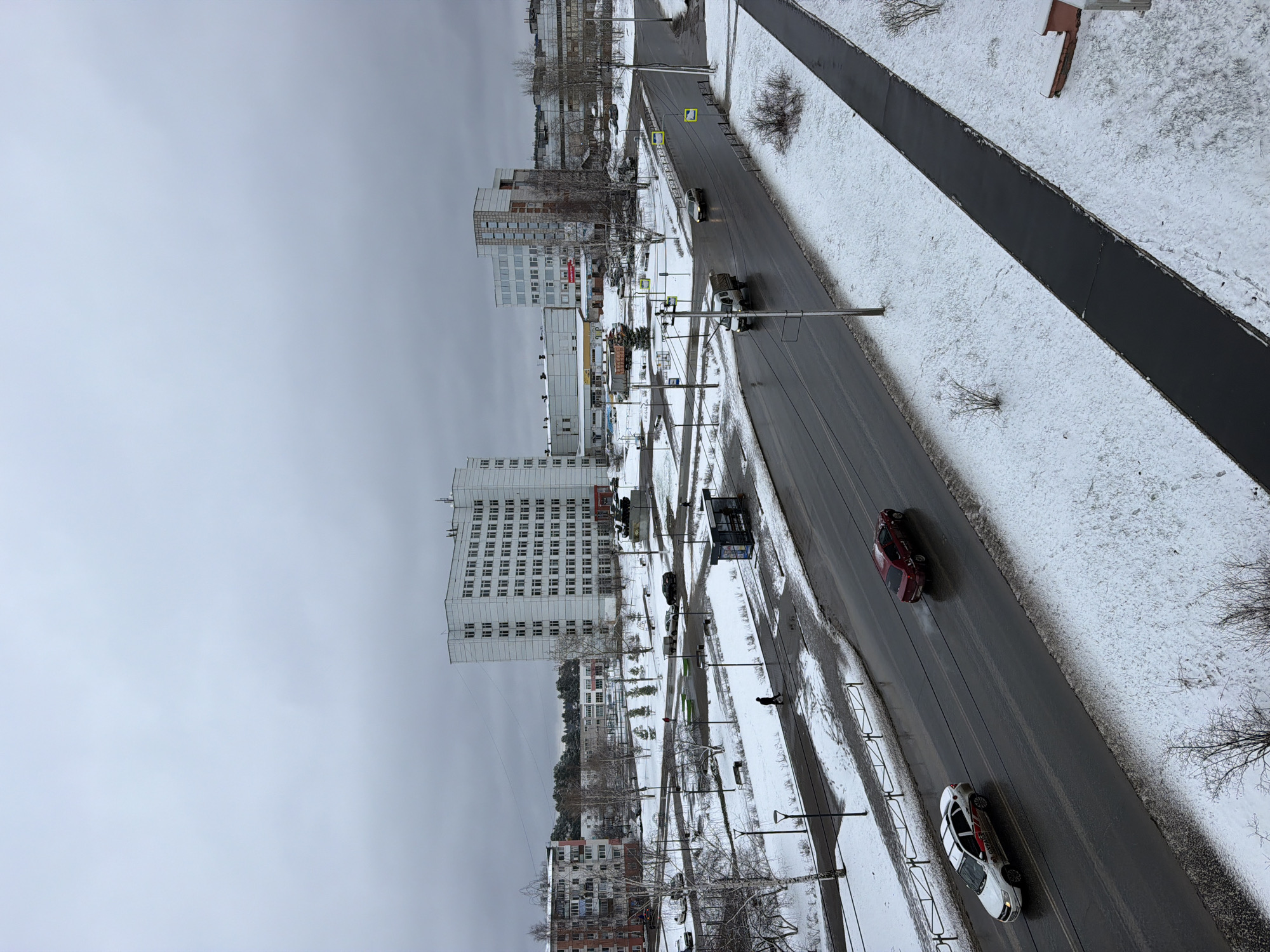 The image presents an elevated perspective of a street scene in the city of Chaikovsky, Russia, during winter. The street is wet, with evidence of melting snow. Buildings of varying heights line the street. Cars are seen driving on the road. The surrounding areas show signs of snow cover on sidewalks and grassy areas. The sky is overcast.