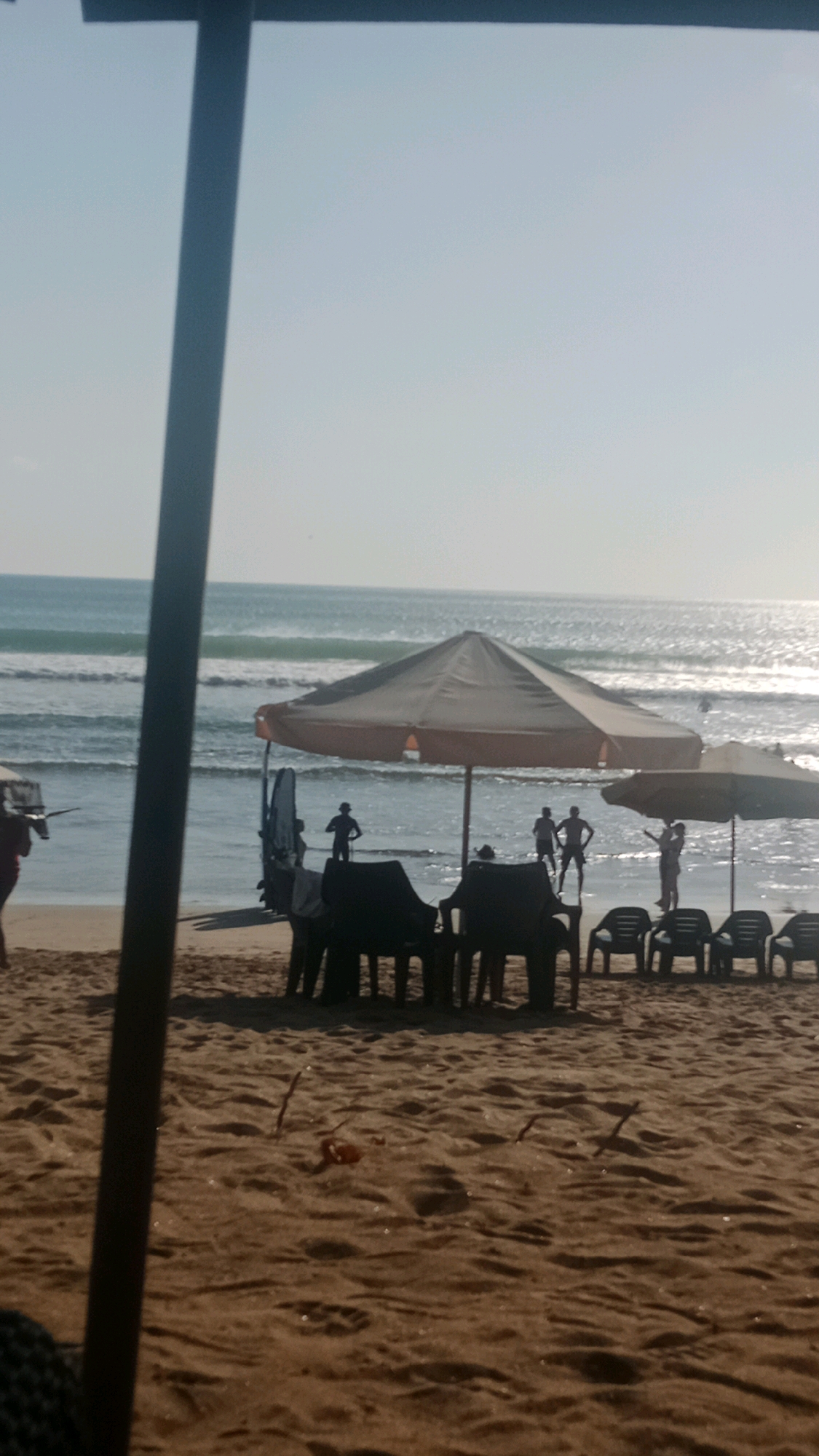 A silhouetted image captures a beach scene in Kuta, Indonesia. In the foreground, dark sand is textured with footprints and debris. A dark, vertical pole partially obscures the view on the left. Further back, several black plastic chairs are arranged on the sand. Behind the chairs, two large beach umbrellas stand, one a light beige and the other a lighter, possibly white, color. In the shallow water beyond the umbrellas, several people are visible as silhouettes. One person stands near a surfboard. The ocean exhibits rolling waves under a clear, pale sky. The scene is bathed in the soft light of what appears to be early morning or late afternoon.