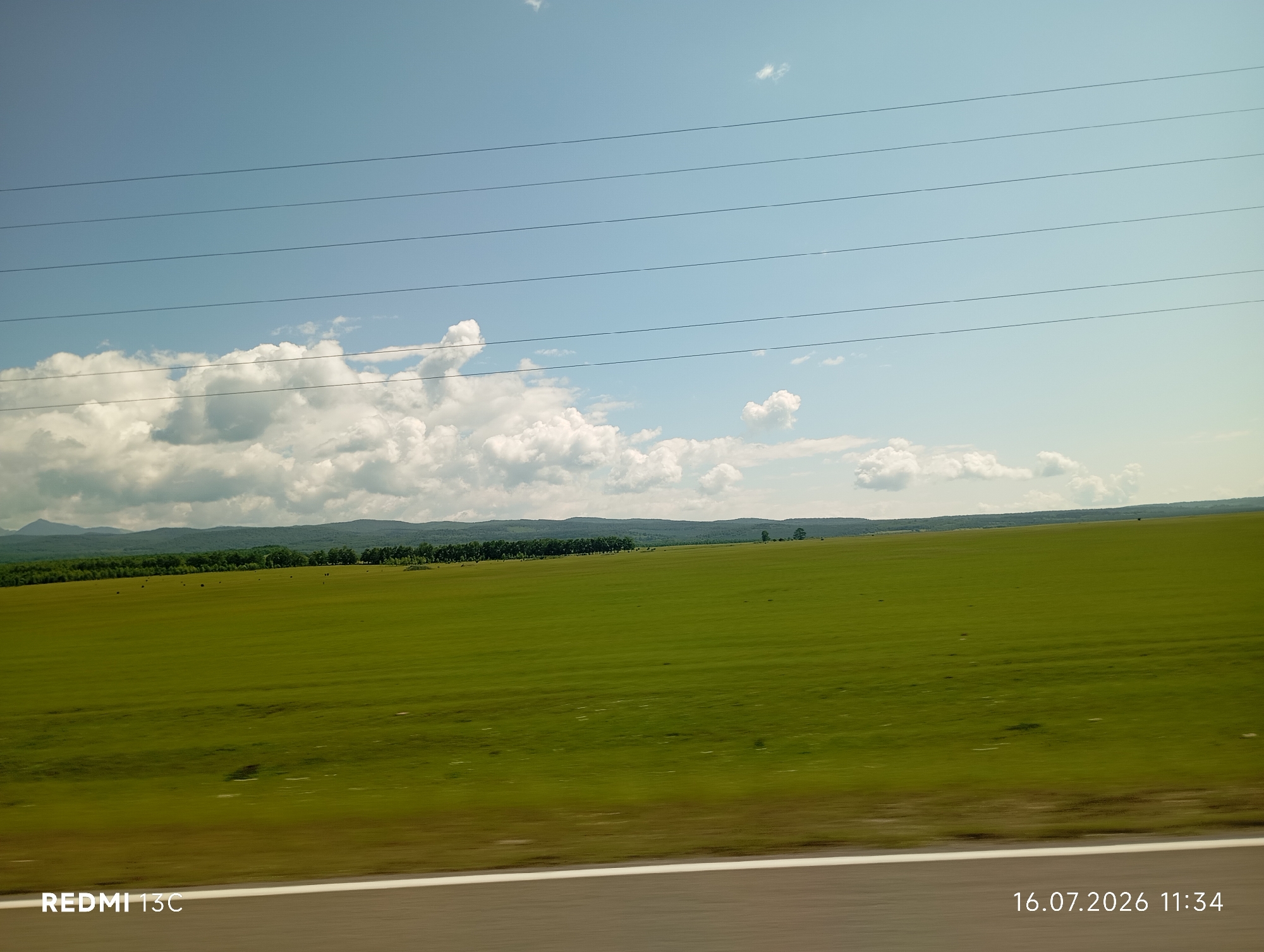 The image depicts a vast, rolling green field under a bright blue sky with scattered white clouds. A line of trees forms a distant horizon, beyond which rolling hills are visible. Several power lines stretch horizontally across the sky. In the foreground, a white stripe marks the edge of a road. The image is a landscape view from a moving vehicle, as evidenced by the slight motion blur. The bottom left corner displays the text "REDMI 13C", and the bottom right corner shows the date and time "16.07.2026 11:34". The location is identified as the city of Galbai, Russia.