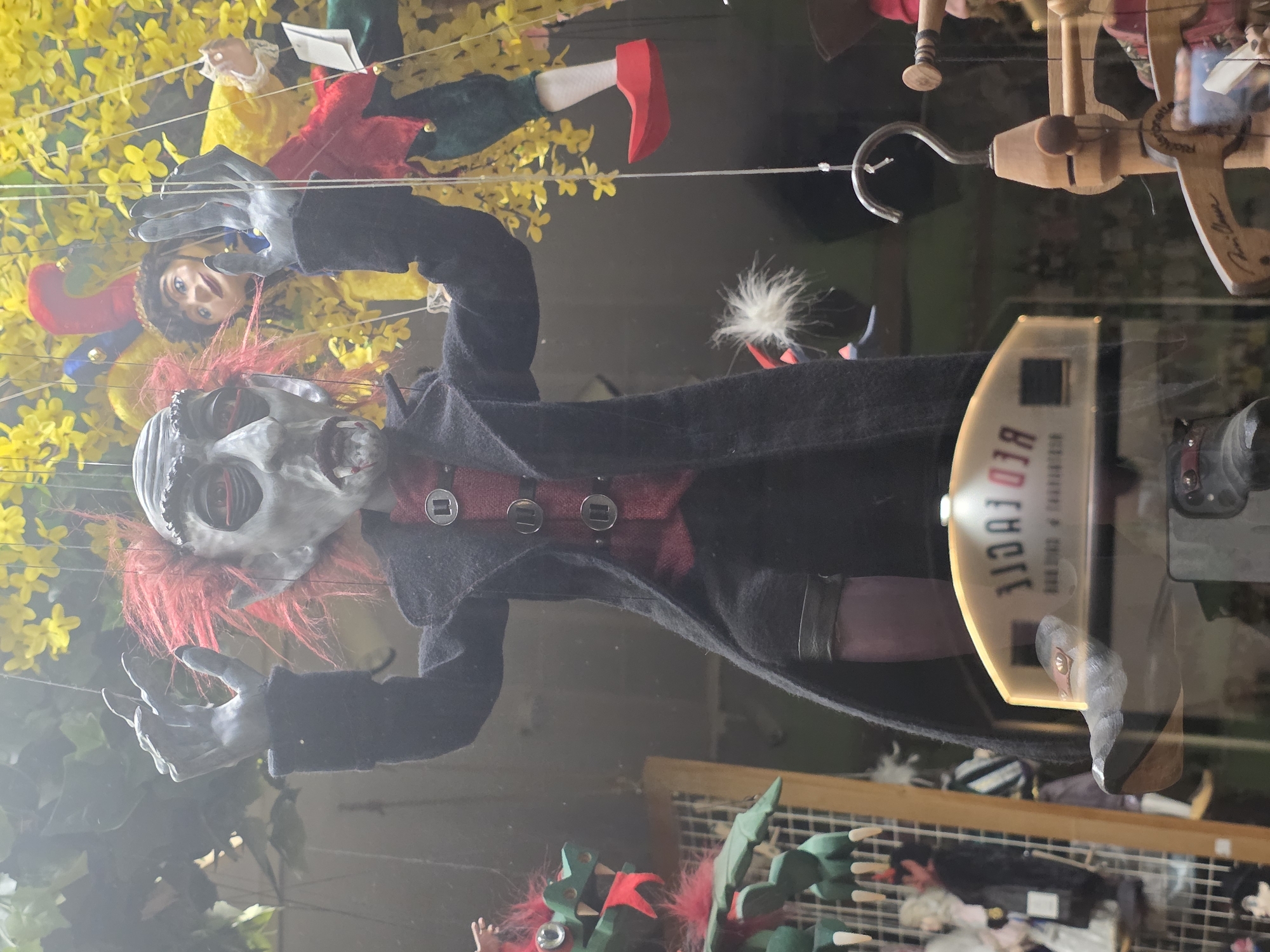 The image displays a collection of hanging puppets, likely marionettes, in what appears to be a shop window or display case. The main subject is a striking puppet with a skeletal or vampiric face, piercing red eyes, and shaggy red hair. It wears a long black coat over a red vest adorned with three silver buckles, and has long, grey, claw-like hands. This puppet is suspended by thin white strings.
Above and to the left of the central figure, another puppet is visible. This one resembles a jester with a multi-colored (red and yellow) hat featuring bells, a yellow top, and a white ruffled collar. Its face is more human-like, with blue eyes. Further to the top right, parts of another puppet are visible, dressed in green and red with red pointed shoes, suggesting an elf or another jester.
To the right of the central puppet, a wooden control bar for a marionette is hanging, featuring a metal hook. Partially visible text on this control bar reads "RED FOCCIE". The background features yellow artificial flowers, creating a somewhat ethereal or whimsical setting, contrasting with the macabre central puppet. The entire scene is viewed through glass, as evidenced by reflections, including a subtle reflection of a phone or camera in the lower right. In the bottom right corner, a green wooden puppet resembling a creature with spiky features and red accents is partially visible. The overall impression is one of an artistic or novelty display of puppets.