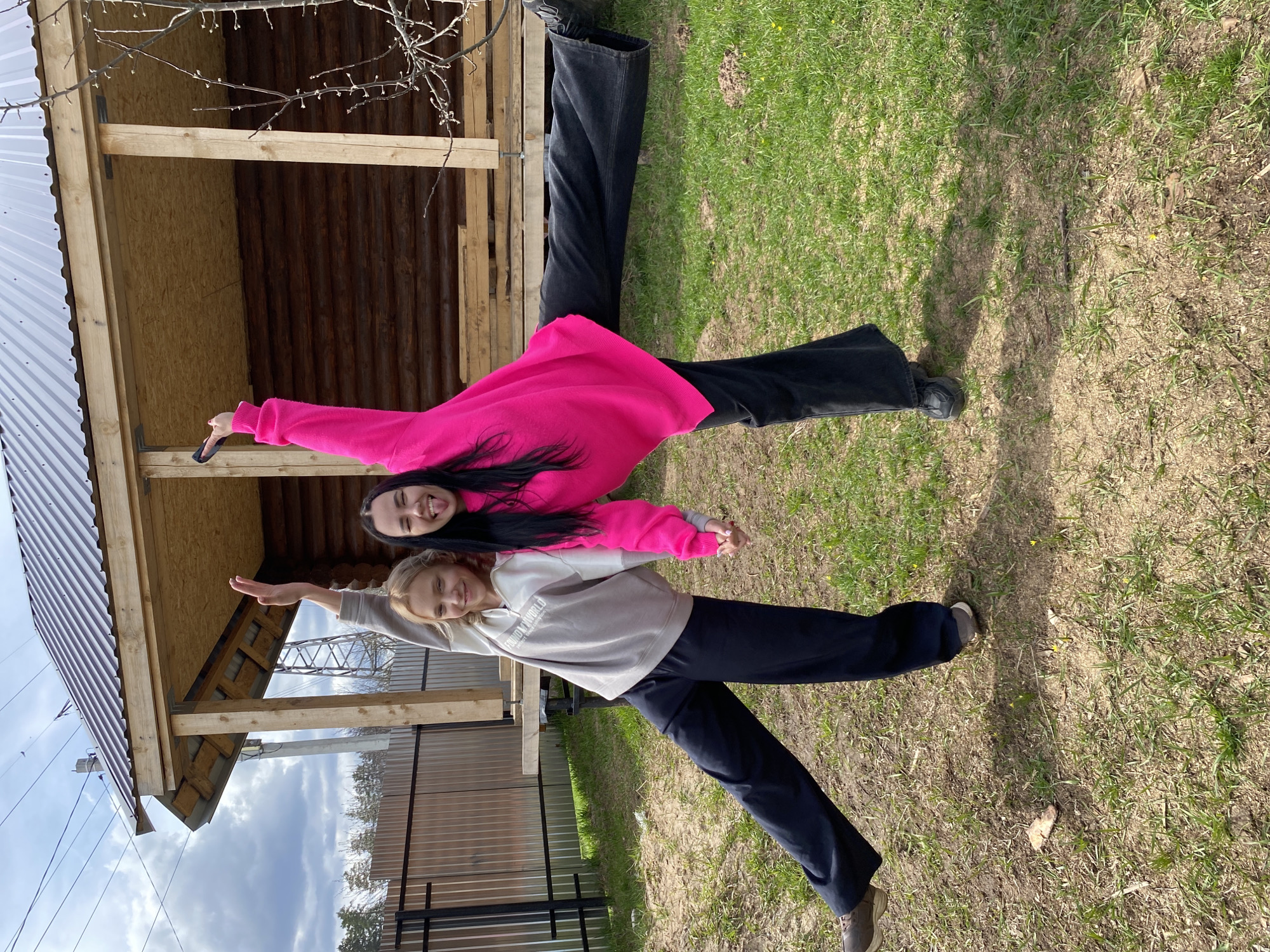 Two young women are captured posing playfully outdoors in what appears to be Chaikovsky, Russia, on a sunny day. They stand side-by-side, holding hands with their arms outstretched, both smiling broadly at the camera, conveying happiness and a cheerful mood.
The woman on the left (when correctly oriented) has long dark hair and wears a bright pink long-sleeved top paired with black wide-legged pants. She holds a dark object, possibly a phone, in her left hand. The woman on the right has blonde hair and is dressed in a light gray long-sleeved hoodie and dark blue pants. The text "FRIDAY.WORLD" is partially visible on her hoodie.
The setting includes a rustic wooden structure, likely a shed or outbuilding, with a corrugated metal roof visible on the left side of the frame. The ground beneath them is covered in green grass with some drier patches and small yellow flowers, indicating early spring. Shadows are cast across the grass, suggesting bright sunlight. Bare tree branches with visible buds are in the upper part of the image. The background features a blue sky with scattered clouds above a metal fence and utility poles.
