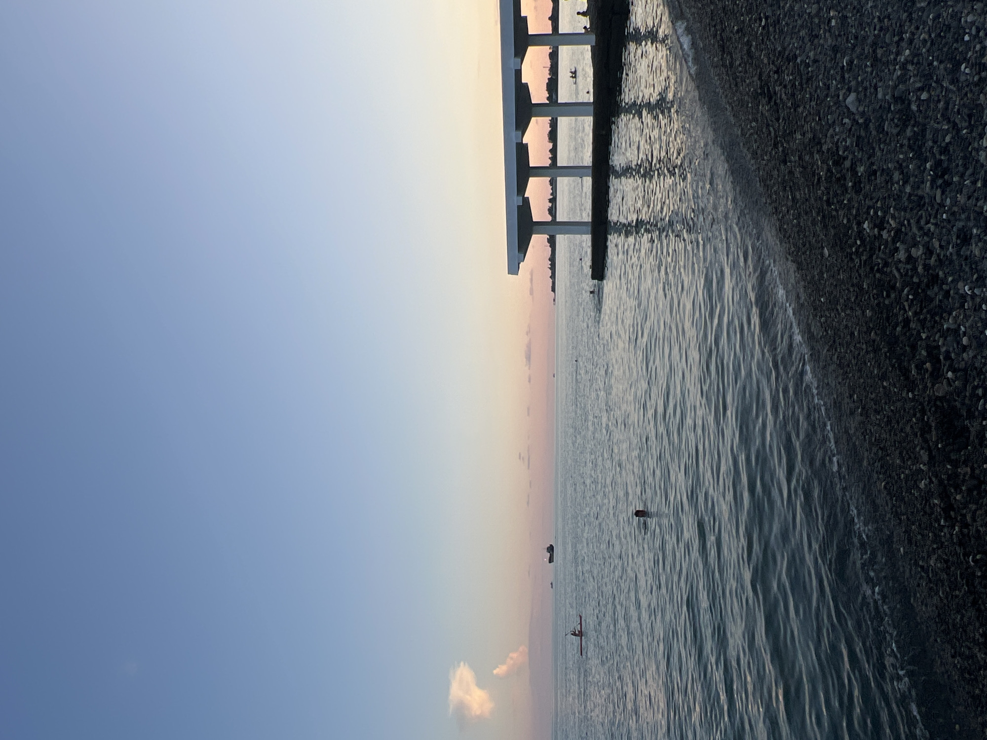 The image depicts a calm sea with a pebble beach in the foreground. The water is a deep blue, with gentle ripples on the surface. Several people are visible in the water; one person is swimming, their head just above the surface, while others are further out, some appearing to be in kayaks. In the distance, a large ship is sailing on the horizon. To the right, a modern white structure with multiple pillars extends over the water, casting reflections on the sea. The sky is a soft gradient of blue, transitioning to a pale pink and orange near the horizon, suggesting either sunrise or sunset. The overall scene conveys a peaceful atmosphere of a coastal leisure area, likely in Sokhumi, Abkhazia.