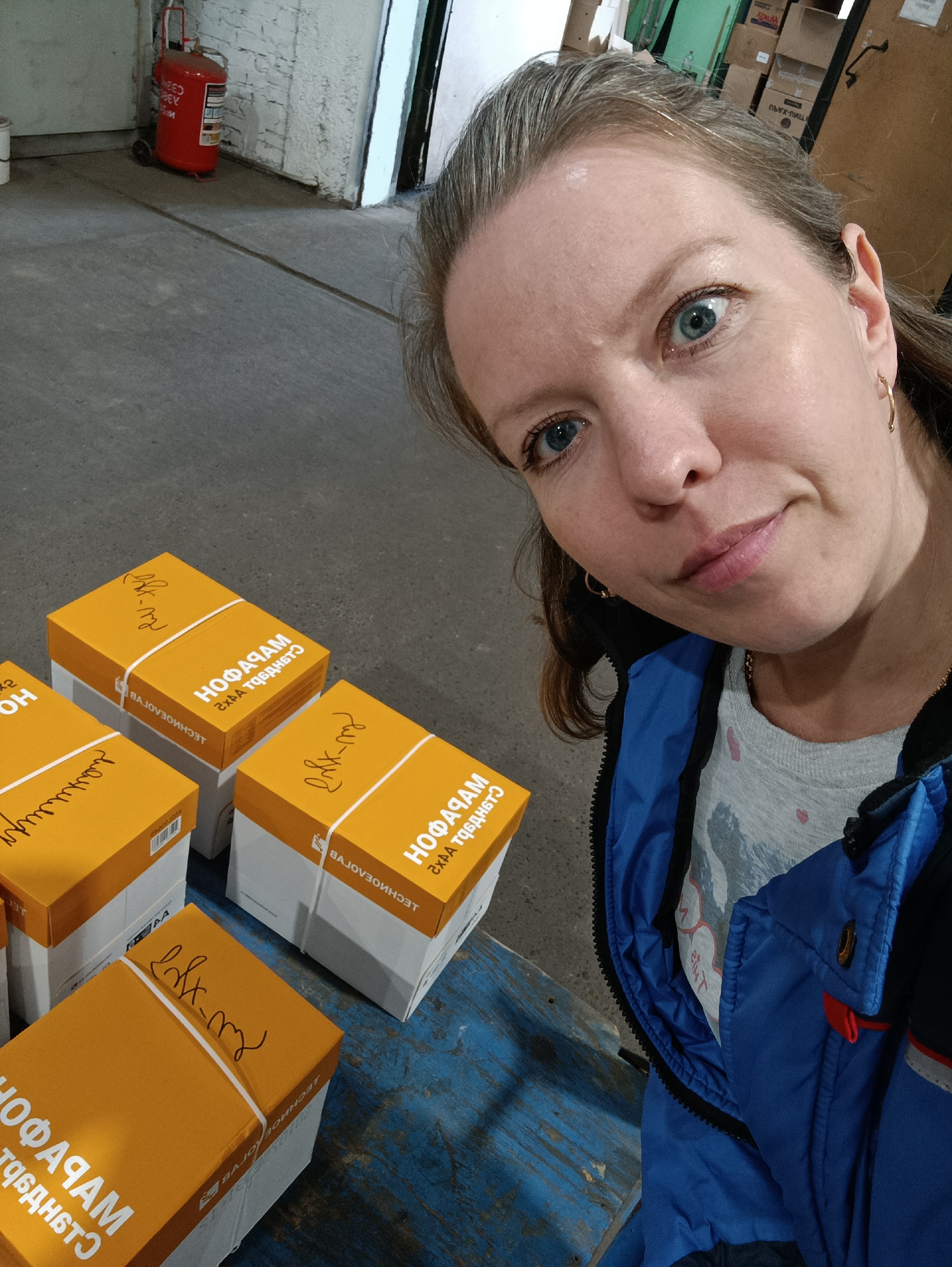 A woman with blue eyes is taking a selfie in what appears to be a warehouse or storage area. She is wearing a blue jacket over a light gray shirt. In front of her are several yellow boxes with white labels with the text "HO OФАЧАМ СХАА ТЬДЬНБТЭ" and "ДЕСНИОЕЛОГУВ" as well as additional writing in black marker, that is in reverse orientation. A red fire extinguisher stands in the background on the left. Behind the woman are stacks of cardboard boxes. The location could be Chaikovsky, Russia, given that the text on the boxes includes Russian words. The floor is a combination of gray concrete and blue-painted wood.