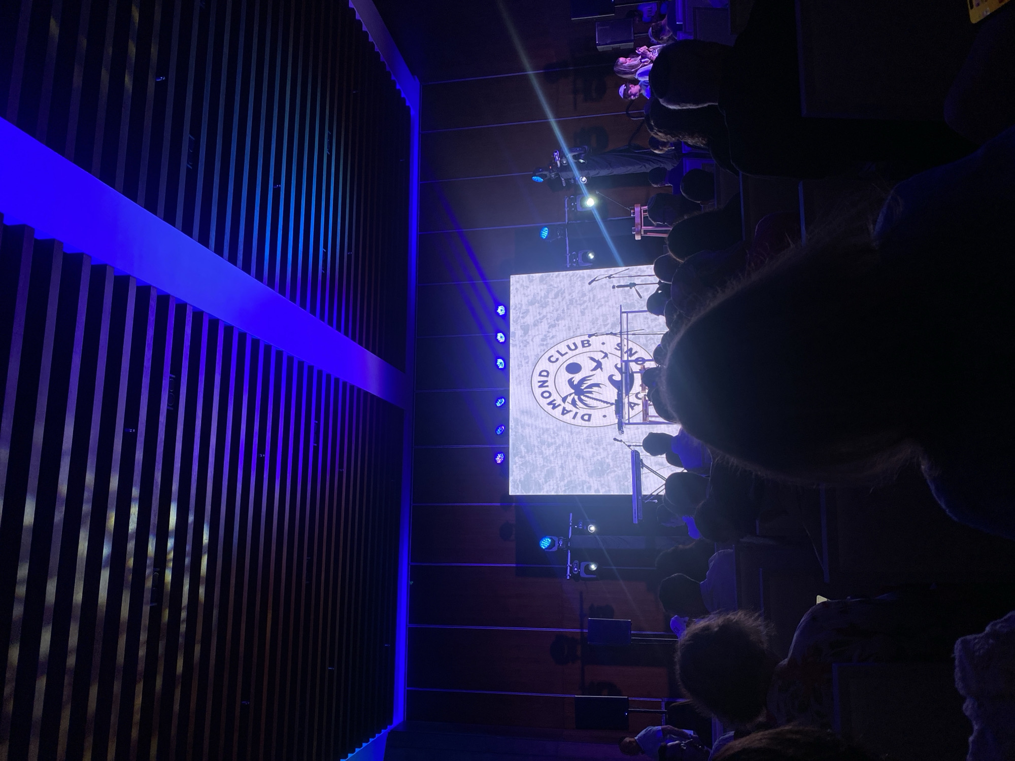 An audience is watching a performance on a stage at the Diamond Club in Marina Maroma, Mexico. A large screen displays the club's logo behind the stage, which is illuminated by stage lights. The interior has modern design elements, including slatted wood walls accented with blue lighting.