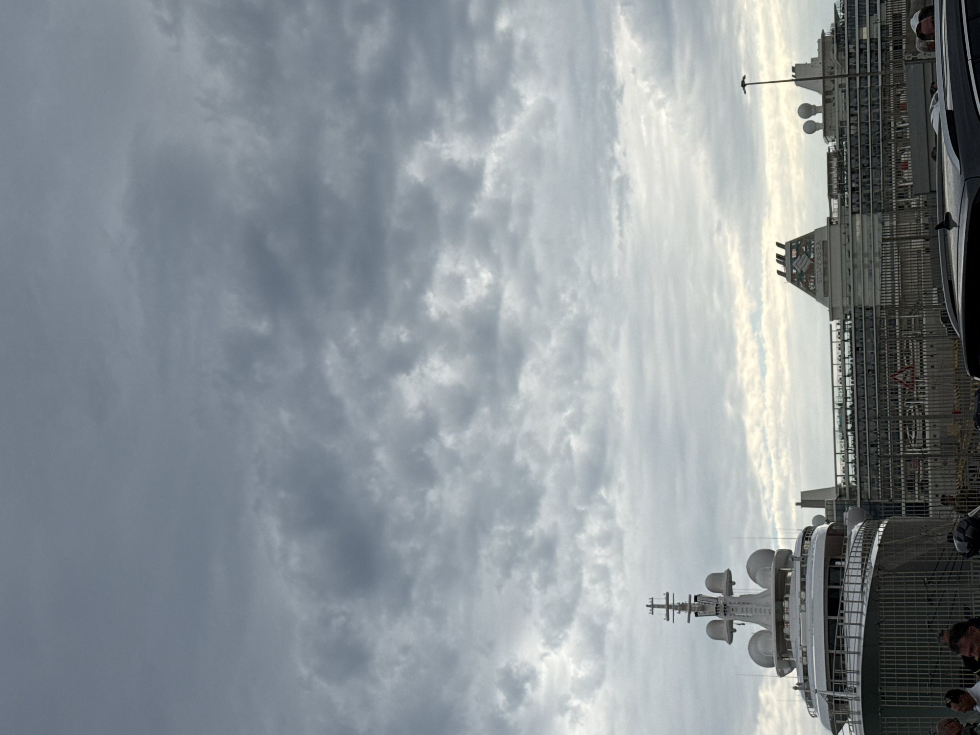 The image shows a partial view of the city of Civitavecchia, Italy, under a cloudy sky. The primary focus is a large cruise ship or yacht docked in the harbor. The ship has multiple levels and a prominent radar or communications mast with several dome-shaped structures. Adjacent to the ship, there is a waterfront area with fences and some infrastructure, and part of a car is visible. The image also captures the surrounding buildings and structures of the city, including a tower with "SILVERSEA" written vertically on it. There are people visible standing in front of the ship, indicating activity on the docks.