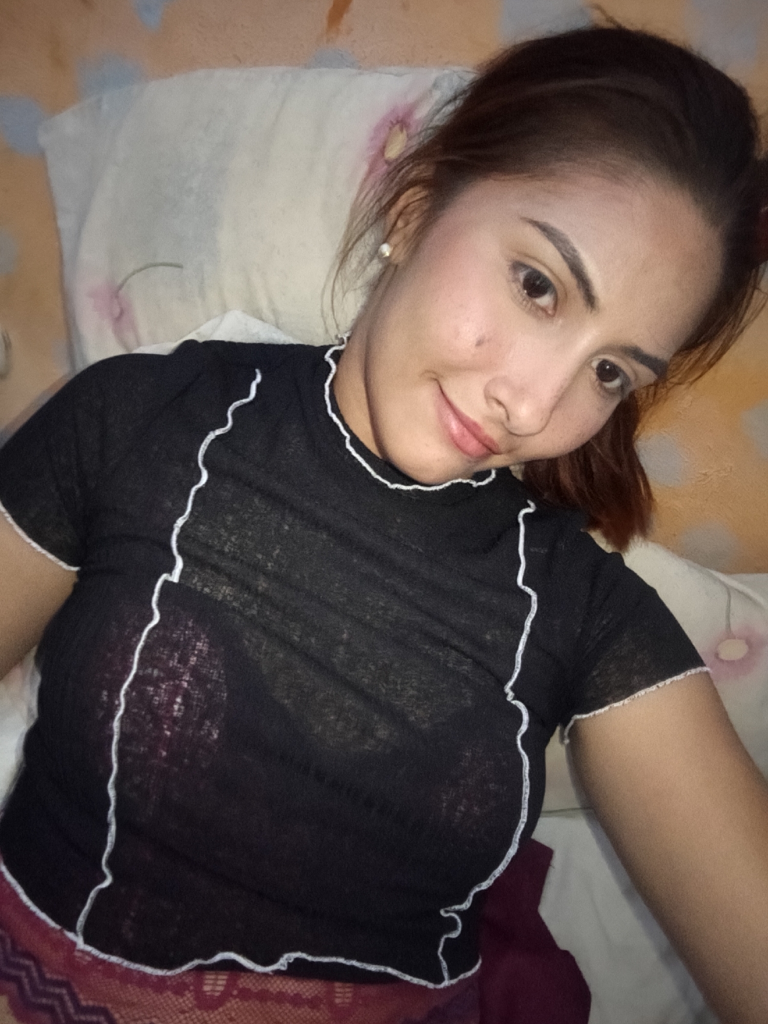 A young adult female is depicted in a high-angle selfie, taken indoors in what is stated to be the city of Juan Griego, Venezuela. She is lying down, supported by pillows. The subject has dark brown hair, pulled back, and a slight, closed-mouth smile. Her eyes are dark, and she wears subtle facial makeup, including a small mole on her left cheek and light-colored stud earrings. She is dressed in a black, short-sleeved, semi-sheer top with distinctive white wavy stitching along the neckline, shoulders, and side seams, and trim on the sleeve cuffs and hem. A dark red or maroon undergarment is visible beneath the sheer top. A portion of a patterned, reddish-maroon lower garment is also visible at the bottom of the frame. The background features light-colored pillows, one with a faint pink stain, resting against patterned bedding in tones of peach, light orange, and light blue.