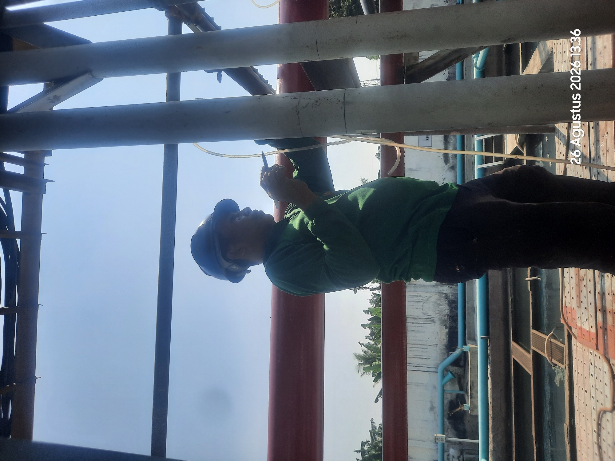 A man wearing a hard hat and green long-sleeved shirt is working on a construction site. He is standing between two large pipes, holding a tool and appearing to be making an adjustment. The background shows a clear sky, some industrial structures, and vegetation. A date and time stamp, "26 Agustus 2026 13.36", is visible in the lower right corner. The location is identified as Gending, Indonesia.