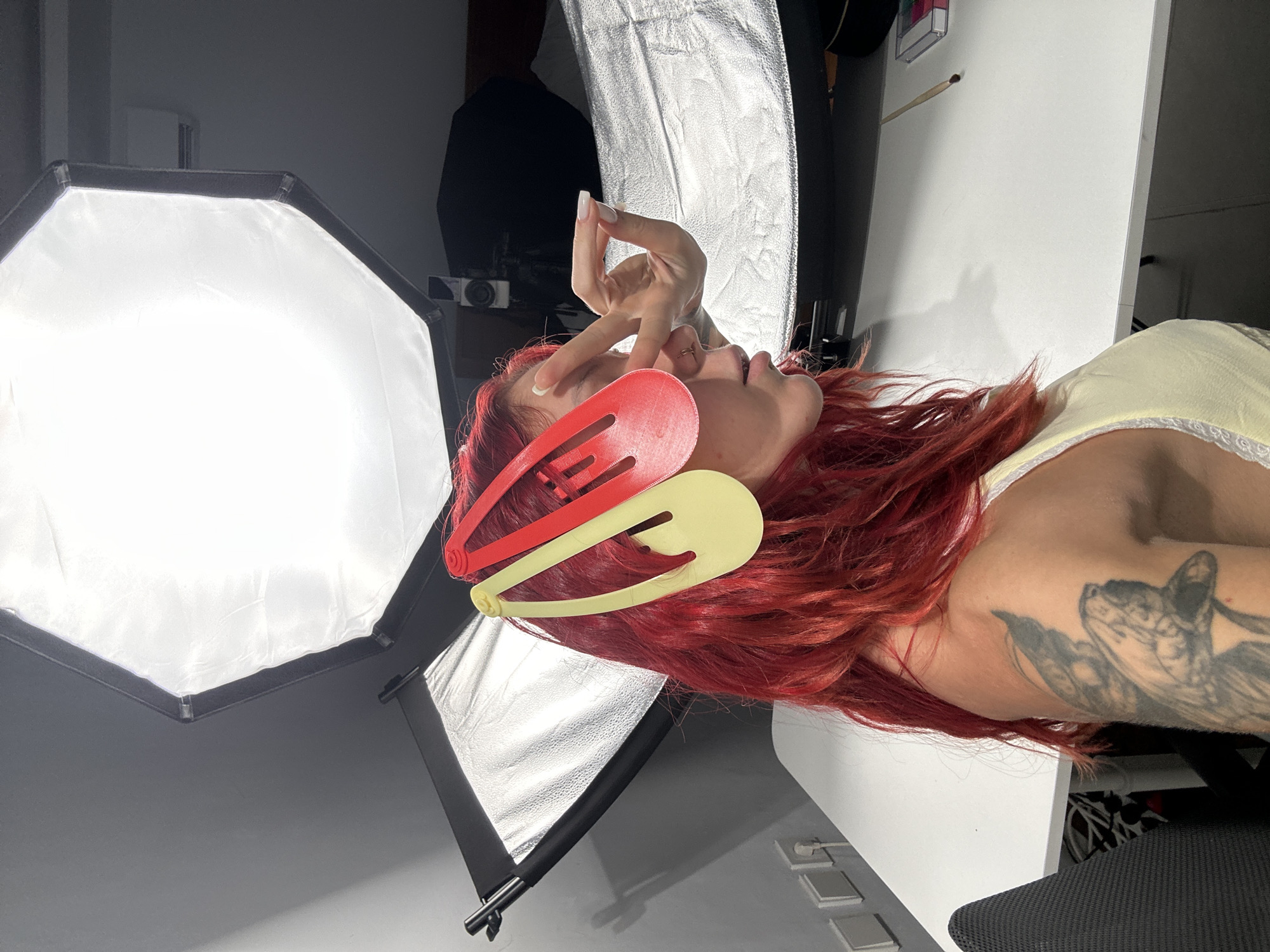 In North Kuta, Indonesia, a woman with vibrant red hair is positioned in a studio setting, likely for a photoshoot. She is wearing a light yellow top with white lace trim. Two large, colorful hair clips, one red and one yellow, are attached to her hair, partially obscuring her face. Her left hand is raised to her face, with her index finger and thumb touching her right eye, creating a playful or artistic gesture. A tattoo is visible on her left arm. The background features professional lighting equipment, including a large octagonal softbox and a rectangular softbox with a silver reflective surface. A white table is situated in front of the woman, with a small palette and a brush on its surface. Electrical outlets are visible on the grey wall behind the table.
