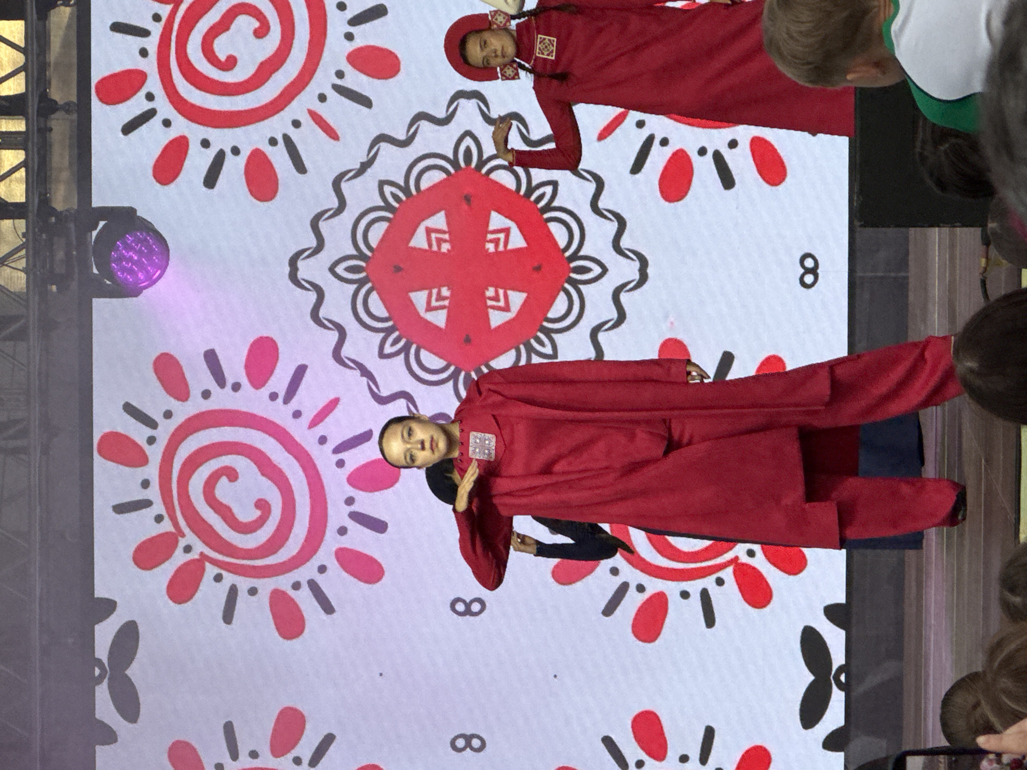 In Chaikovsky, Russia, at an outdoor cultural event, three individuals are on a stage in front of a large screen displaying a folk-art pattern. The screen features large red and pink sun-like motifs with black accents, interspersed with black numbers "8". The foremost subject is a young woman wearing a long, flowing red coat over a red top and pants. She has her left arm raised, with her hand near her chest, and her right arm extended slightly. Behind her to the left, another individual, partially obscured, is also wearing red. To the right, a second young woman stands, dressed in a red outfit with a red hat and pigtails. She is holding her left hand up, gesturing towards the screen. The stage is wooden, and in the foreground, the heads of audience members are visible. A purple stage light is positioned above and to the left of the main subjects, casting a glow on the screen.