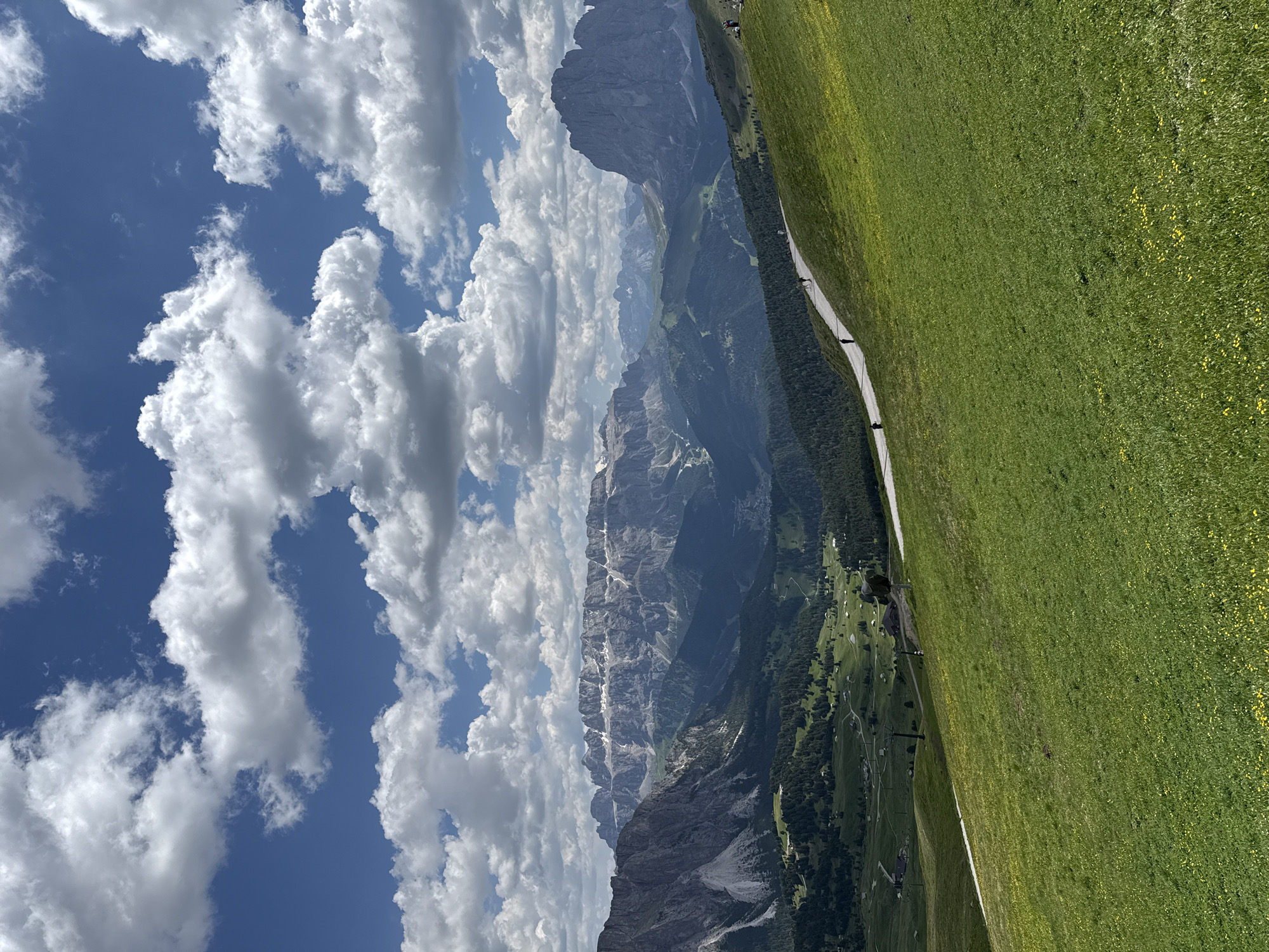 The image shows a panoramic view of the Dolomites mountain range near Santa Cristina Gherdëina, Italy. In the foreground, a lush green meadow slopes gently downwards, dotted with small yellow wildflowers. A paved pathway winds its way up the hill, and several individuals are seen walking or standing along it. In the middle ground, rolling hills covered in dark green forests lead to a valley with scattered buildings and a ski lift. The background is dominated by the majestic, rugged peaks of the Dolomites, some of which have patches of snow. The sky above is a vibrant blue, filled with scattered white, fluffy cumulus clouds. The overall scene depicts a serene mountain landscape on a clear day.