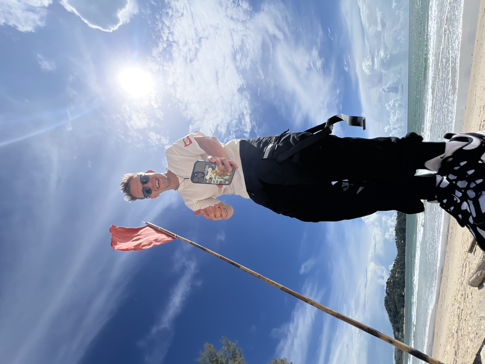 A low-angle, full-body shot captures a man standing on a beach with a red flag and a wooden pole behind him. The man, wearing a white t-shirt, black pants, and white and black sneakers, smiles and gives a thumbs-up while holding a phone. The background features a clear blue sky with scattered clouds, bright sunlight, the ocean with waves, and a sandy beach. A distant green hill with buildings is visible on the horizon. The location is identified as Unknown, Unknown.