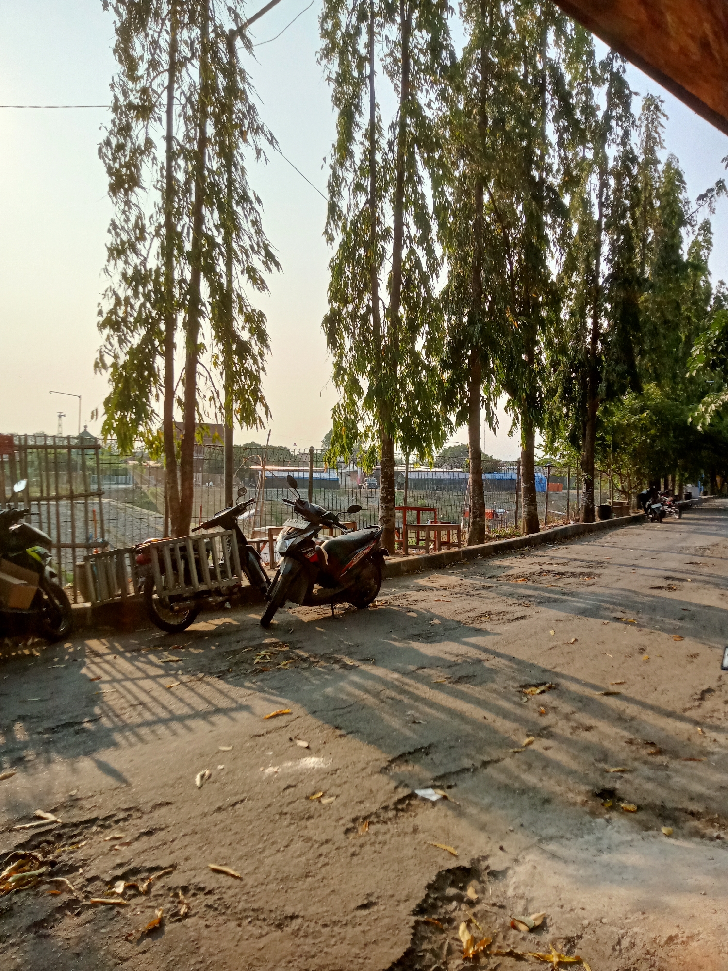In Majan, Indonesia, a paved road runs alongside a row of tall, slender trees with thin trunks and sparse foliage. The trees cast long shadows across the road, indicating it is late afternoon or early morning. Several motorcycles are parked near the trees, including two prominent ones in the foreground. One motorcycle, a dark color with red accents, is parked upright. Beside it, another motorcycle, also dark colored, is leaning on its kickstand. Further back, another motorcycle is visible on the left, its front tire pointing towards the viewer. A metal fence runs along the left side of the road, separating it from an area with what appears to be a body of water. In the background, a street lamp stands tall. The road surface is uneven and appears worn, with debris and fallen leaves scattered across it. The overall atmosphere suggests a quiet, possibly suburban or semi-rural setting.