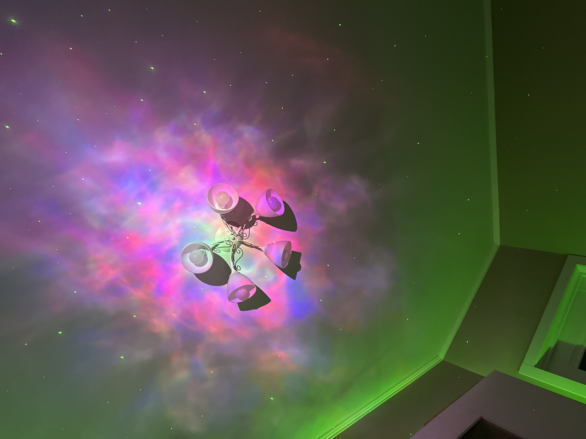 The image shows a low-angle view of a ceiling fan with six lights. The light from the fan projects a colorful nebula-like pattern onto the ceiling, predominantly in hues of pink, purple, and blue. The ceiling itself appears to be painted in shades of green. The image suggests an interior space, with architectural details such as corners and trim visible. The location is Unknown, Unknown.