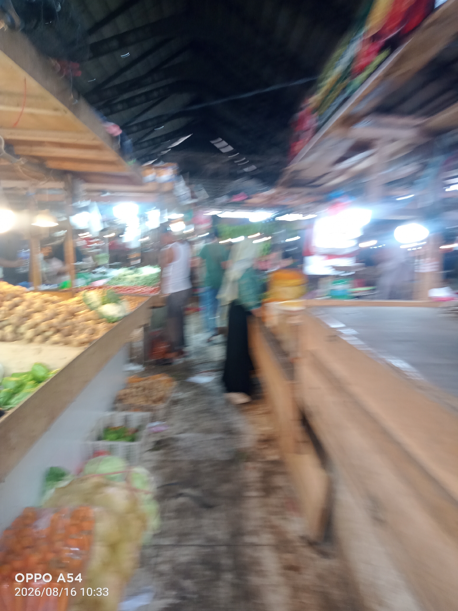 This is a blurred, low-angle interior shot of what appears to be a covered market or food stall. The scene is dominated by stalls laden with produce, including what looks like ginger or potatoes in the foreground left, and other vegetables like cabbage and carrots. Several people are visible, blurred due to motion, some of whom are likely vendors and customers. The lighting appears to be from overhead lamps, casting a warm glow. The overall impression is a busy marketplace with abundant fresh goods. The text overlay "OPPO A54 2026/08/16 10:33" indicates the phone used and the date and time of capture, but provides no information about the specific location within Jambi, Indonesia.