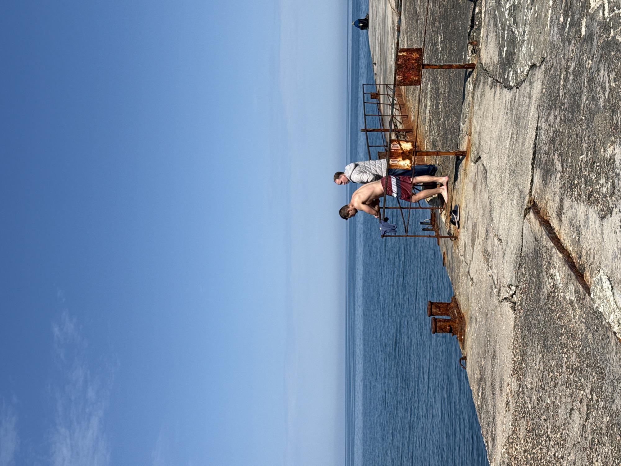 The image depicts two men standing on a concrete structure adjacent to a body of water, possibly the ocean, under a clear blue sky. The concrete structure appears aged, with cracks and rusty metal railings. One man is shirtless and wearing maroon shorts, leaning over the railing to look down at the water. The other man is wearing a light-colored jacket and appears to be watching the first man. The location is Unknown, Unknown.