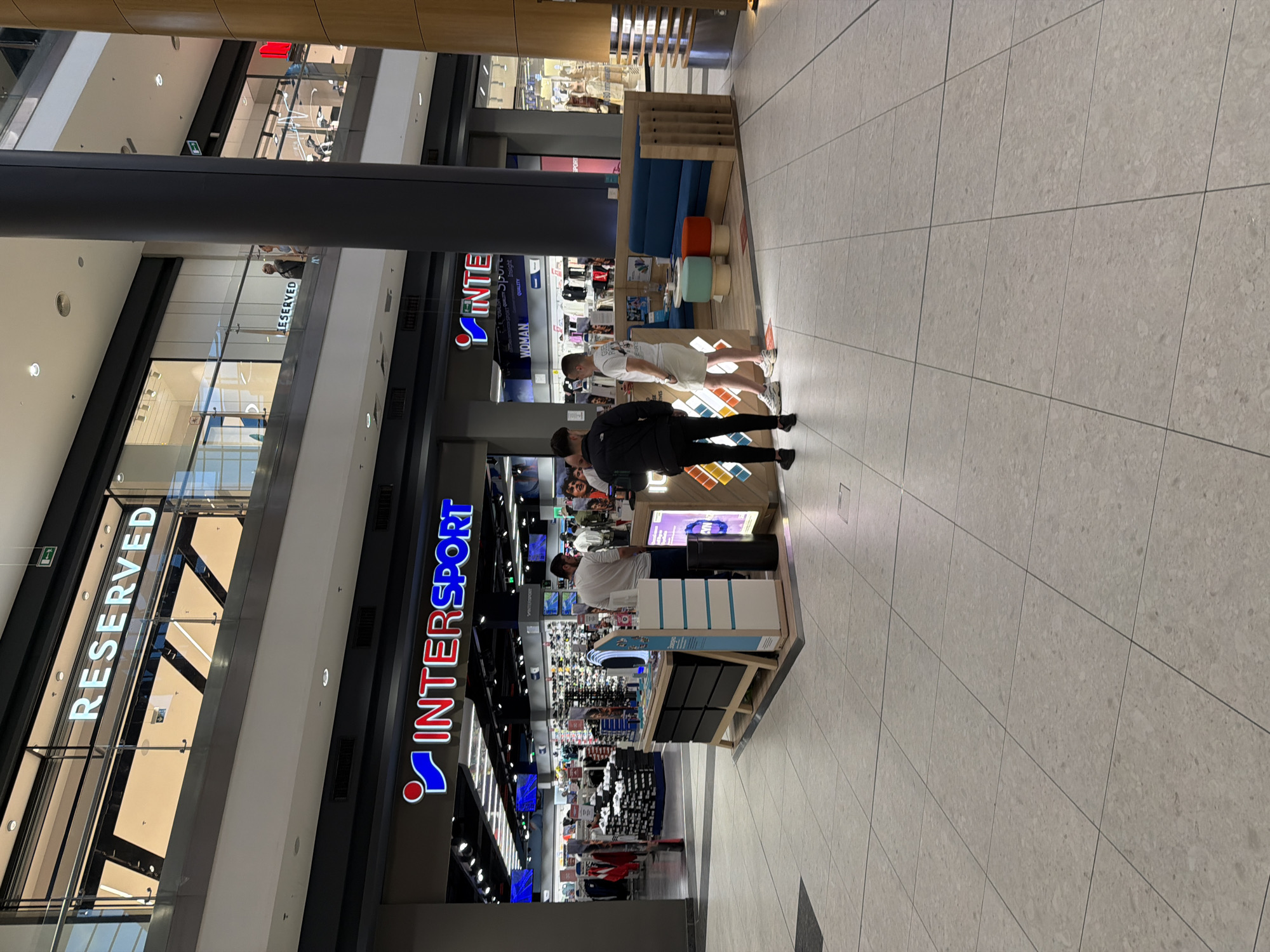 The image shows the interior of a modern shopping mall, potentially in Burgas, Bulgaria, based on the observed signage of international brands present there. The view is from an elevated position looking down.
Foreground: A pedestrian area tiled with large, light-colored square tiles. Two individuals, possibly a father and son, are standing in front of a small information kiosk or display stand. The son is wearing a white t-shirt and light-colored shorts, while the father is in a black jacket. The kiosk itself has a display of electronic items, possibly fitness trackers or smartwatches. An employee in a white shirt attends to them.
Midground: An "Intersport" store prominently displays its logo in red and blue. The store appears to be fully stocked with sports equipment. Above the kiosk, there's a lounge area with a blue couch and colorful stools.
Background: The mall structure is multi-level with glass railings. Another shop, "Reserved," is visible on an upper level. The lighting is bright and modern. The presence of multiple floors and several retail outlets indicates a large, contemporary shopping center.